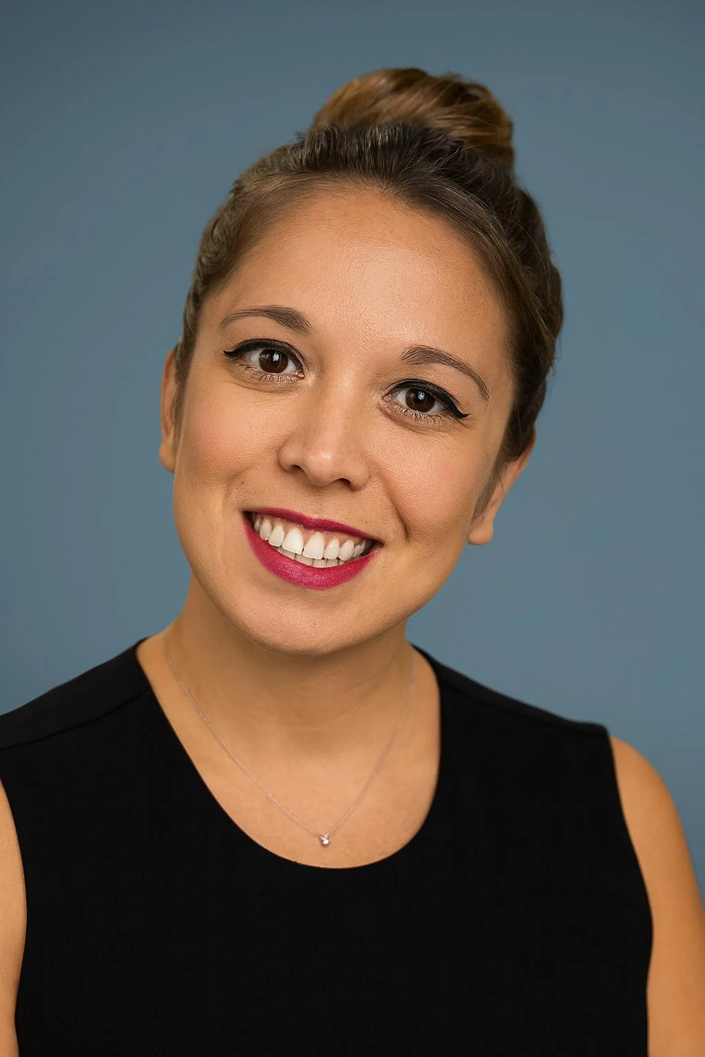 A woman with light brown hair styled in a bun, wearing a black sleeveless top and a silver necklace with a small pendant, smiling against a blue background.