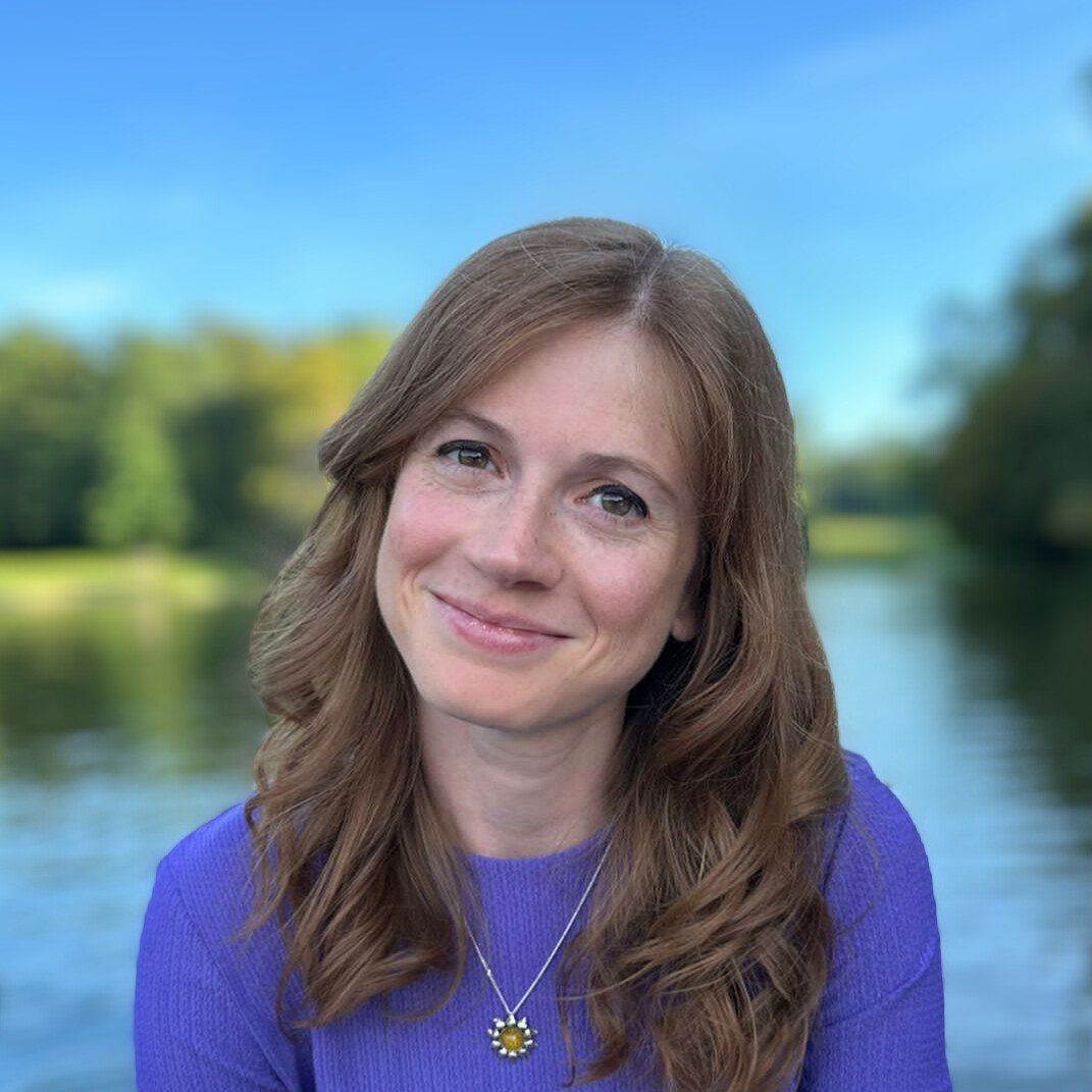 A woman with wavy, shoulder-length brown hair and blue eyes, wearing a purple sweater and a necklace with a flower-shaped pendant, smiling outdoors with a river and trees in the background under a clear blue sky.