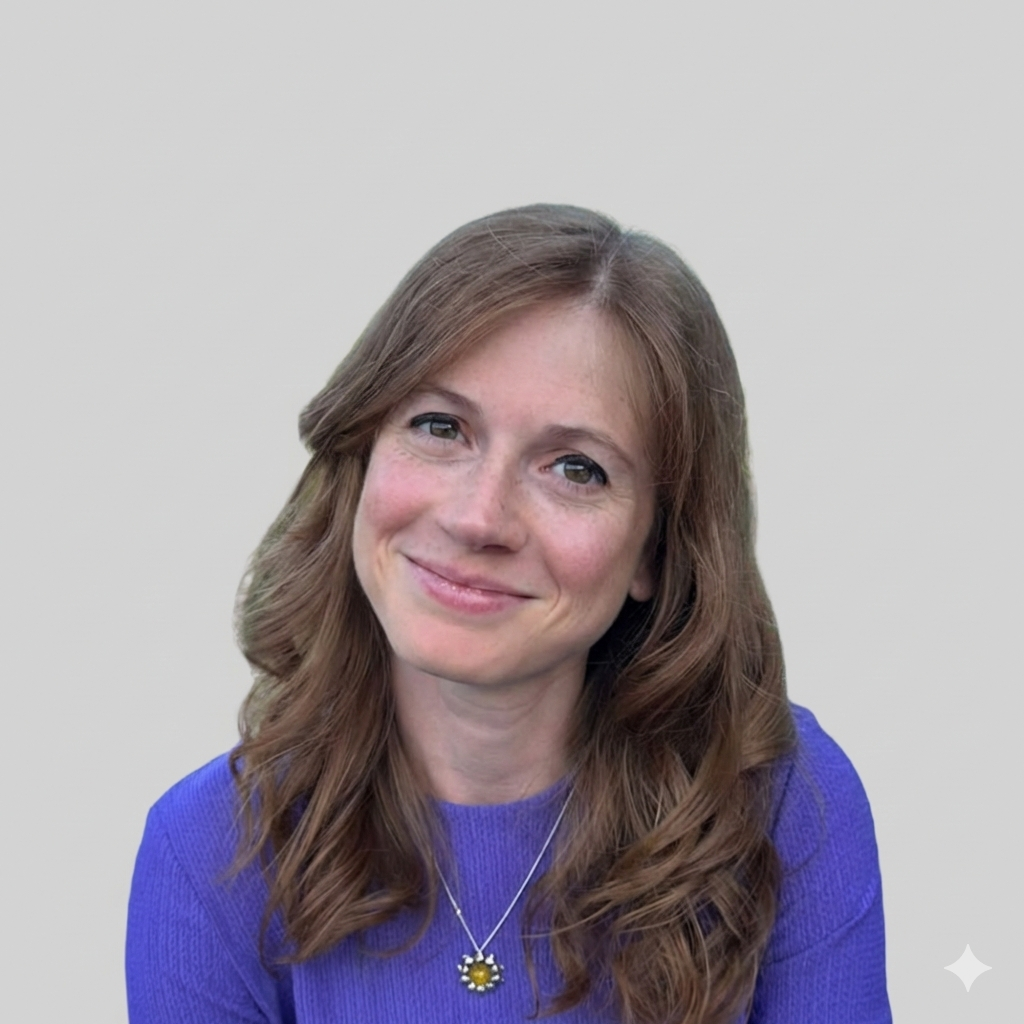 A woman with wavy, shoulder-length brown hair and blue eyes, wearing a purple sweater and a necklace with a flower-shaped pendant, smiling outdoors with a river and trees in the background under a clear blue sky.
