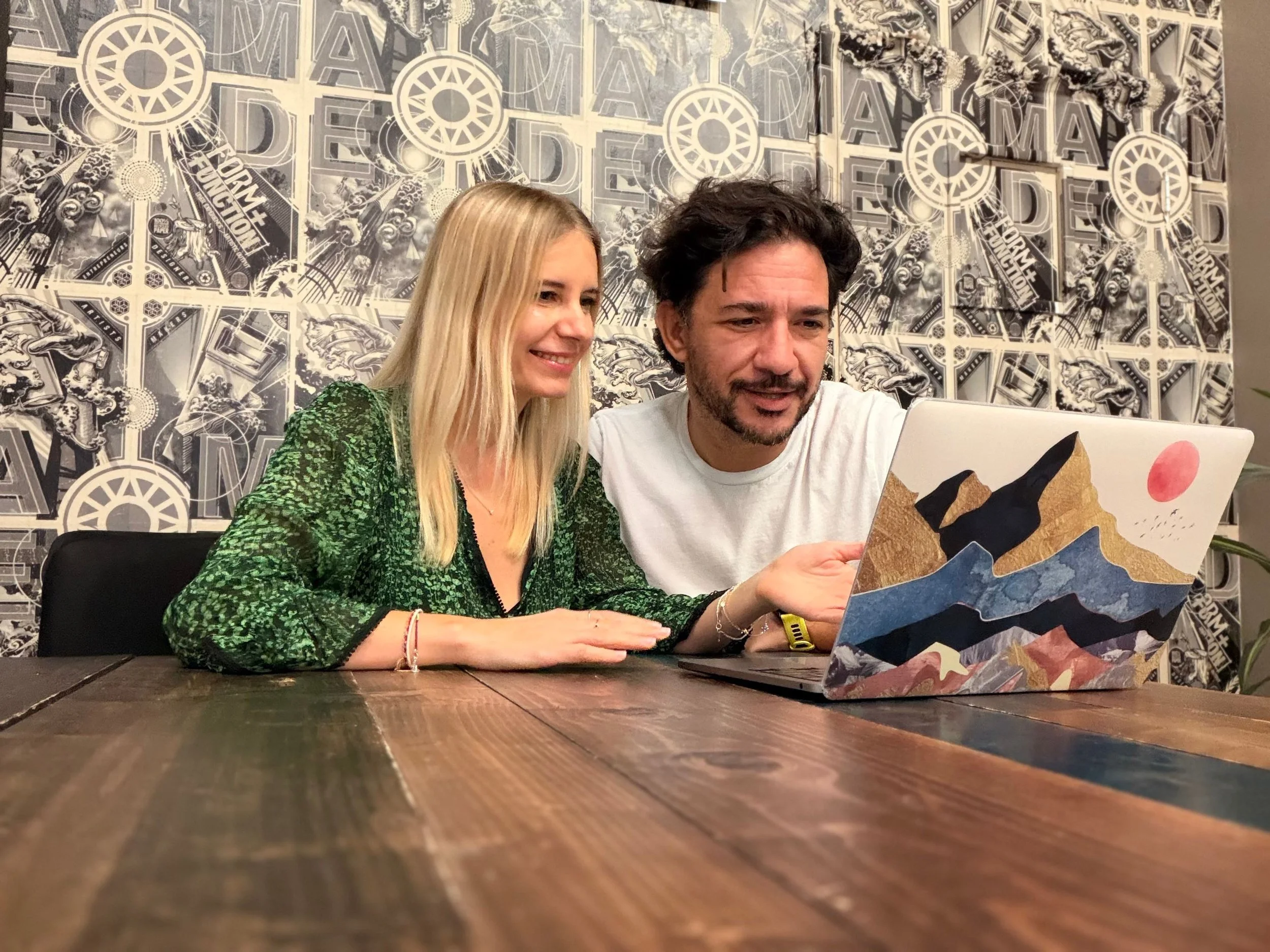 Two people, a woman with blonde hair wearing a green patterned blouse and a man with dark hair and a beard wearing a white shirt, look at a laptop with a colorful mountain landscape sticker, sitting at a wooden table in a decorated room.