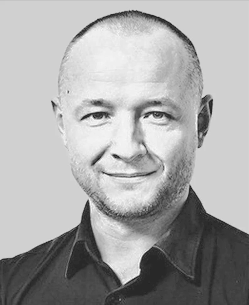 Black and white portrait of a smiling Caucasian man with a shaved head and light beard, wearing a collared shirt against a plain background.