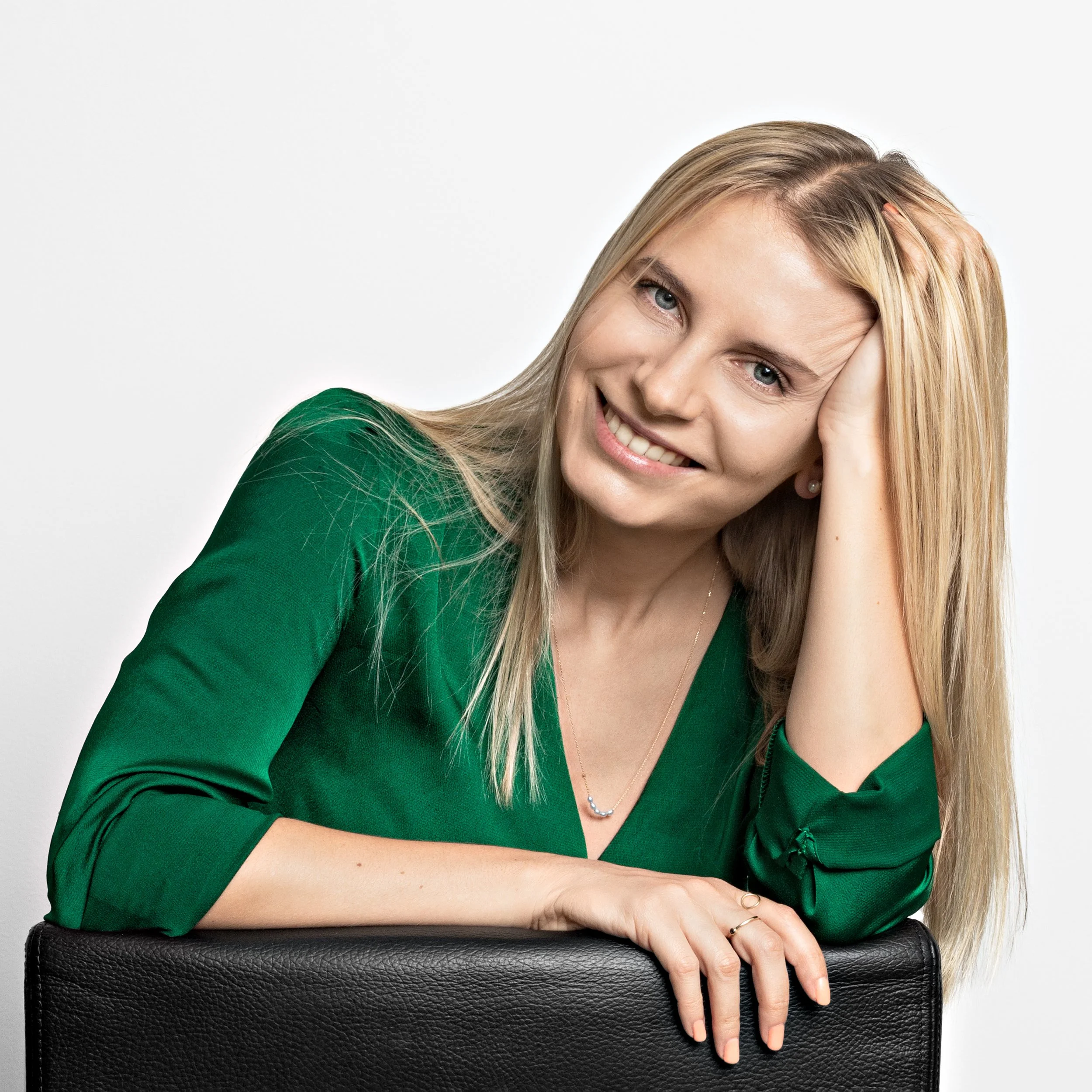 A woman with long blonde hair, wearing a green top, smiling, resting her head on her hand, leaning on a black textured surface against a plain white background.