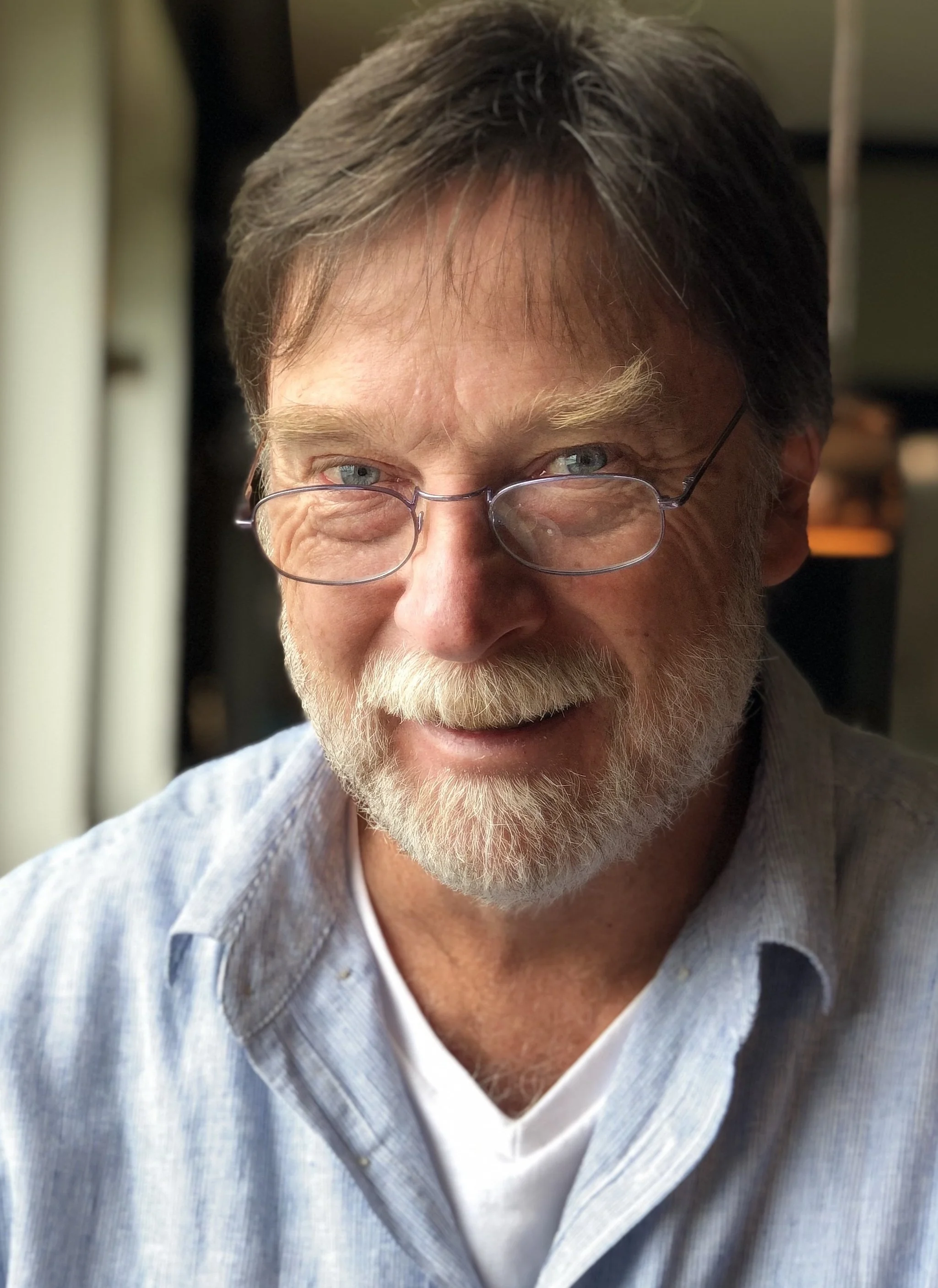 A smiling older man with glasses and a beard, wearing a light-colored shirt, sitting indoors near a window with daylight.