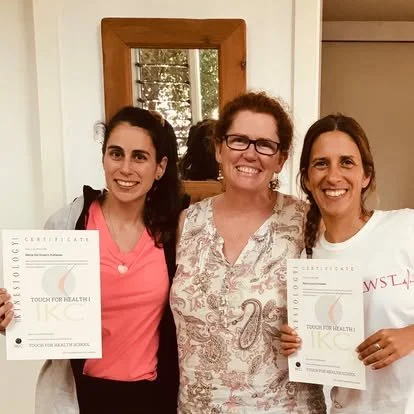 Three women smiling indoors, two holding certificates, with a mirror and window behind them.