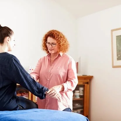 A woman with curly red hair, wearing glasses and a pink shirt, smiling and holding hands with another woman in a dark blue shirt in a bright room.