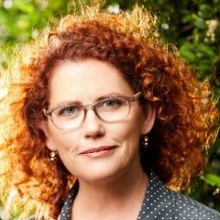 A woman with curly red hair wearing glasses and earrings, outdoors with greenery in the background.