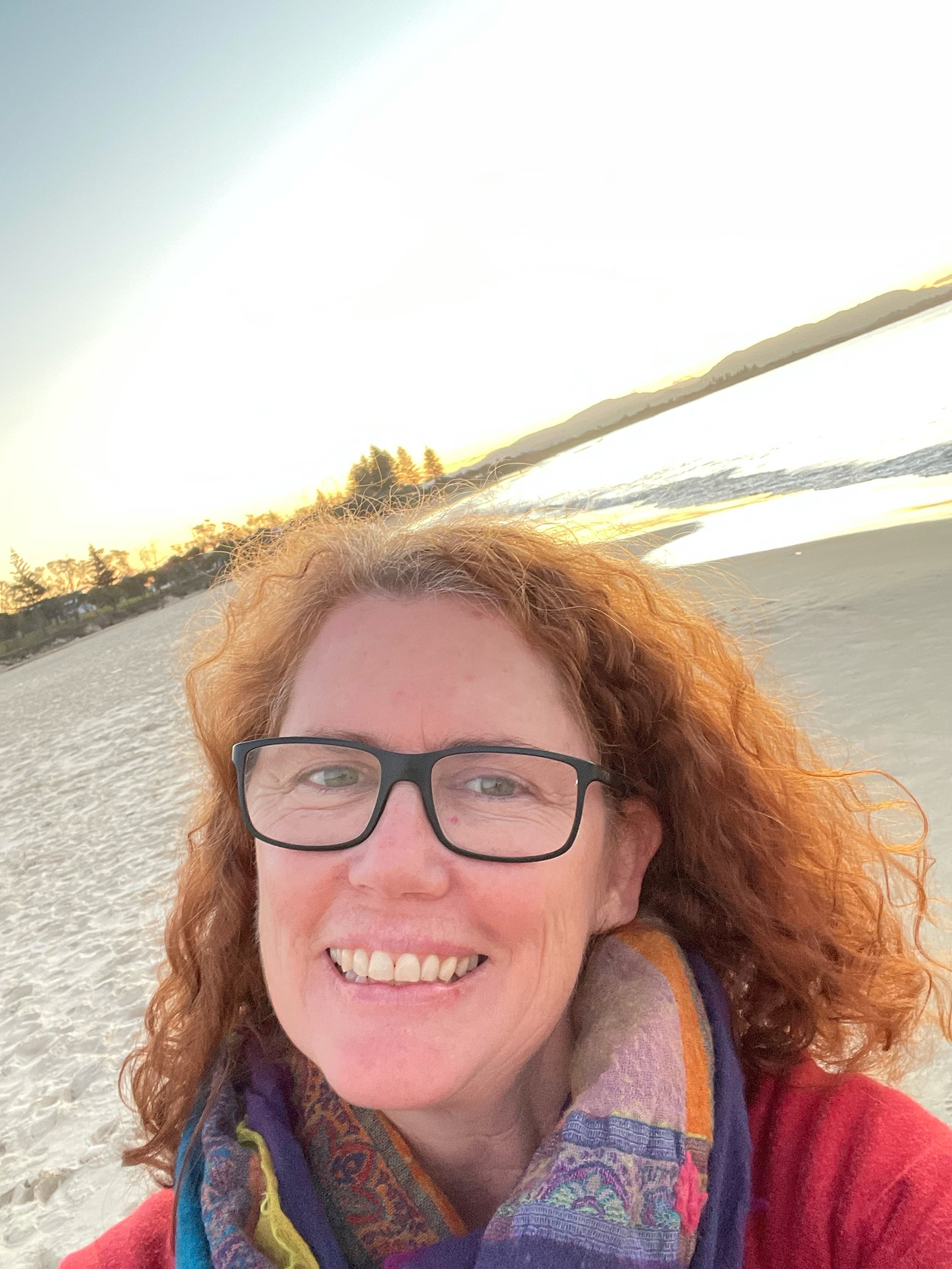 A woman with red curly hair, wearing glasses and a colorful scarf, smiling on a sandy beach at sunset with the ocean and distant hills in the background.