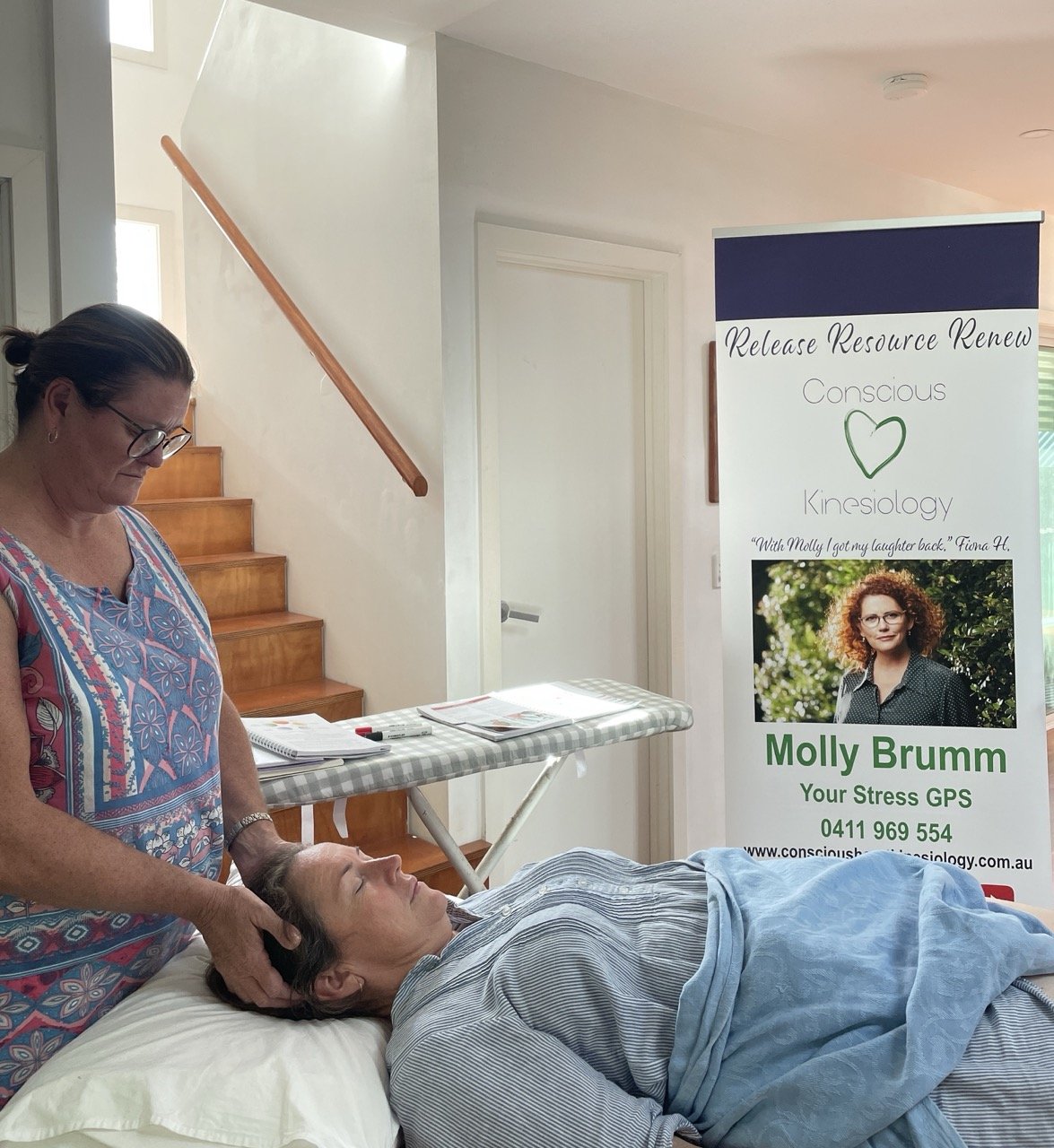 A woman receiving a massage or therapy from a practitioner in a home or clinic setting, with a promotional banner nearby for Molly Brumm, a kinesiologist.