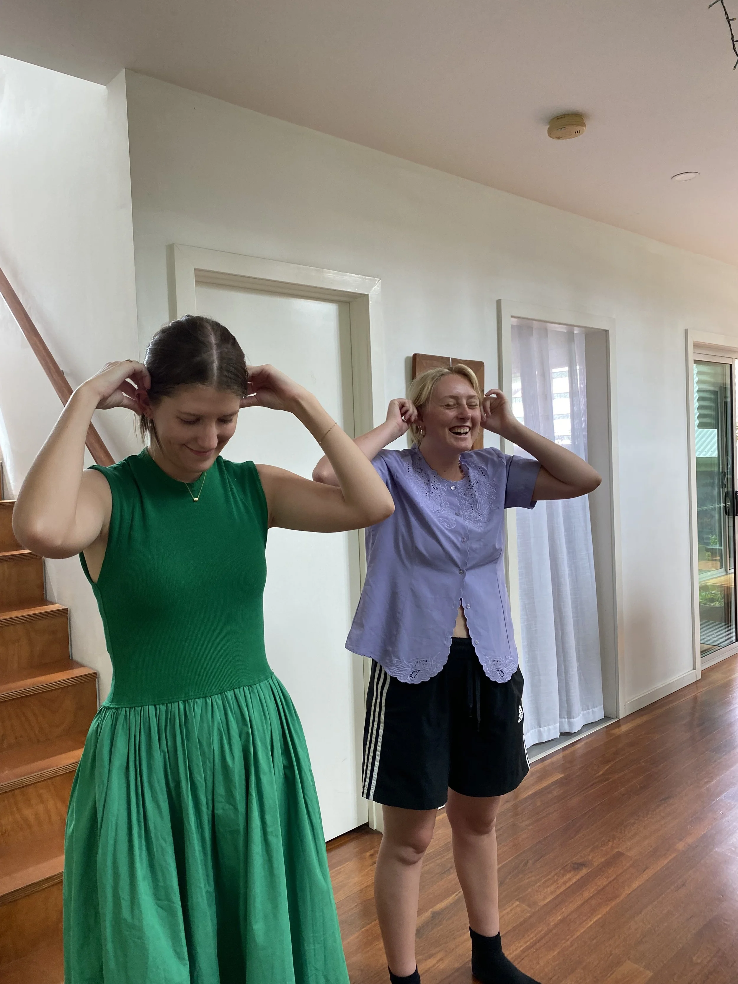 Two women standing indoors, smiling, with hands near their ears, in a room with wooden floors and a staircase in the background.