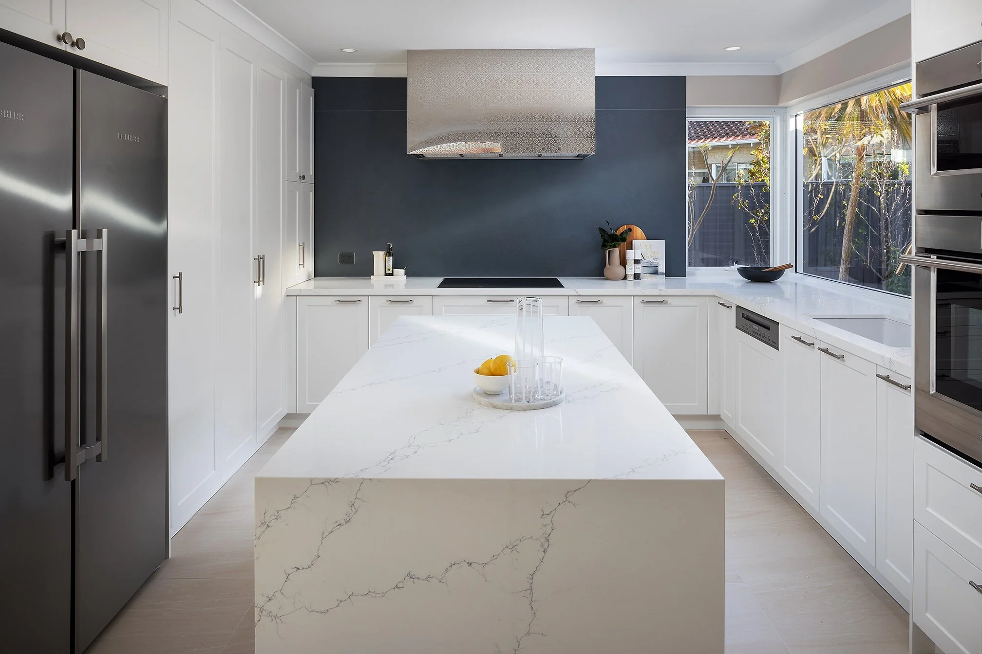 Modern kitchen with white cabinets, a marble island counter, stainless steel refrigerator, built-in dishwasher, and a large window with a view of outdoor trees.