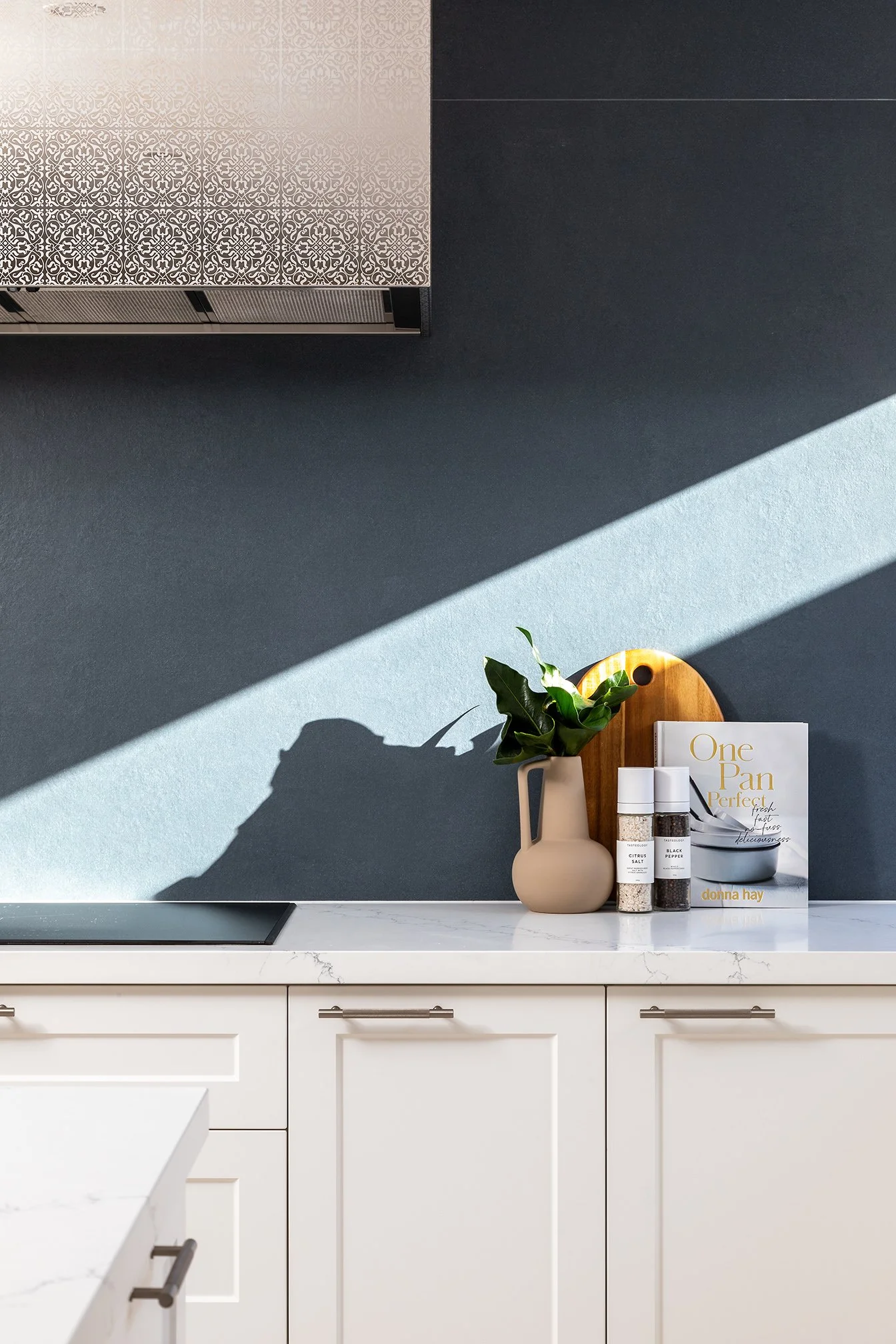 Kitchen counter with a beige vase holding green leaves, a wooden cutting board, two spice bottles, a book titled 'One Pan Perfect' by Donna Hay, and a black bowl on a white marble countertop, with dark blue wall and sunlight casting shadows.