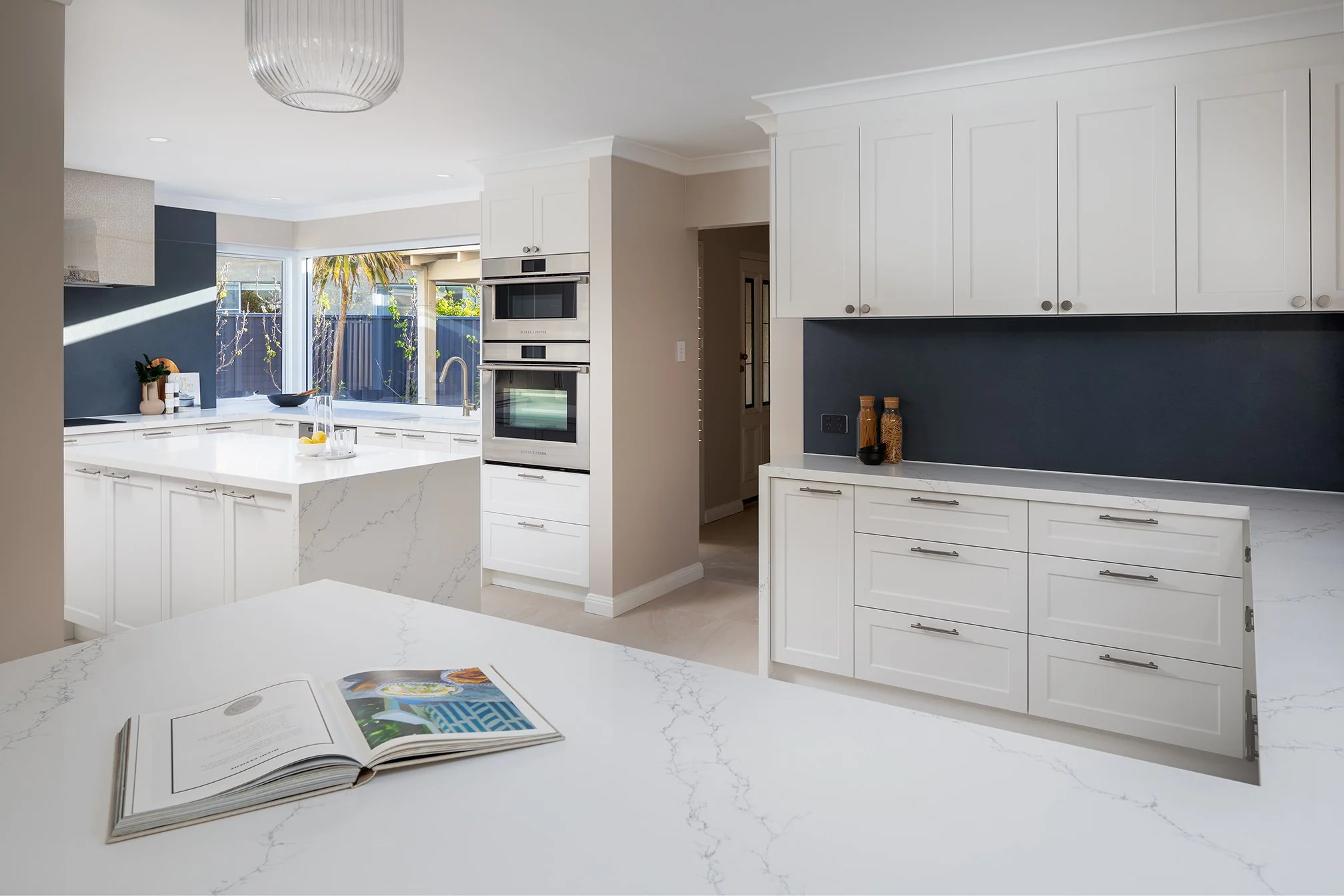 Modern white kitchen with marble countertops, built-in double oven, and large window with a view of palm trees outside.