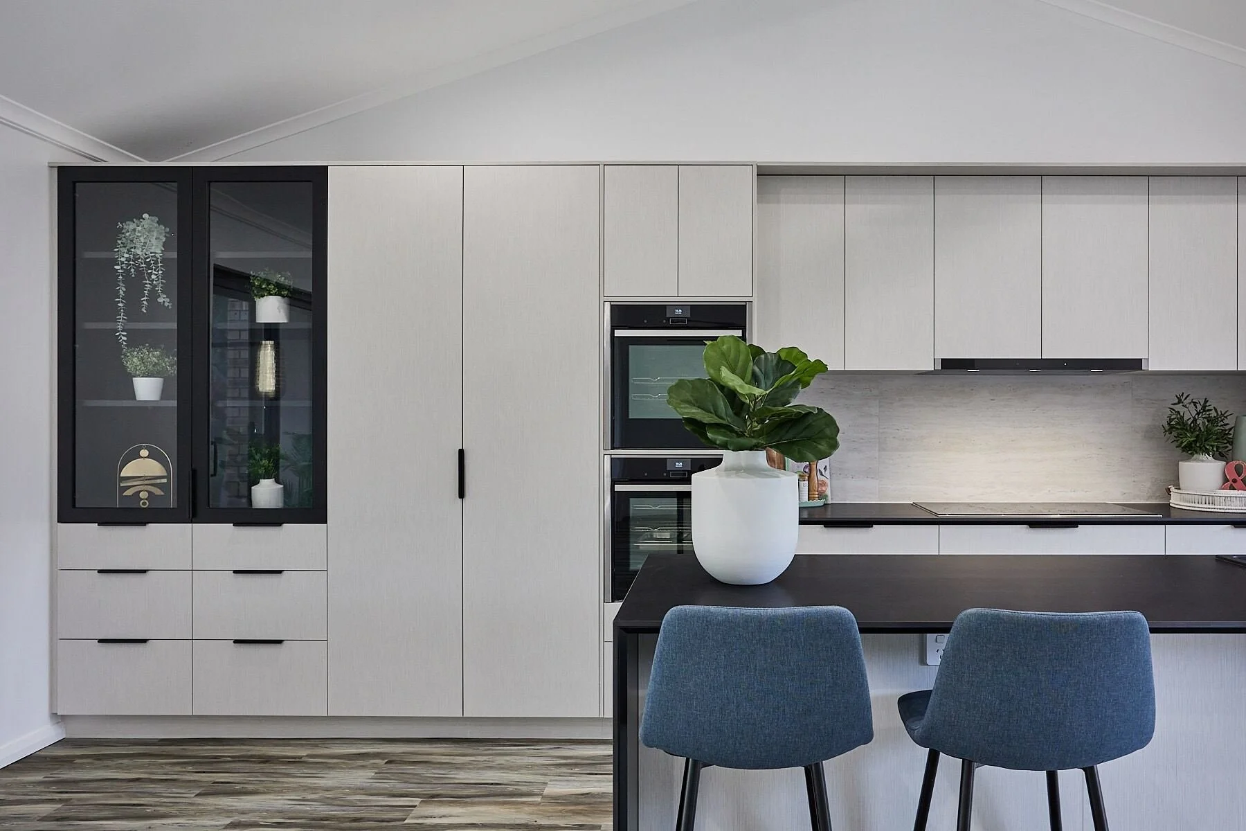 Modern kitchen with light grey cabinetry, built-in ovens, a black countertop, and a small dining area with two blue chairs and a white potted plant.