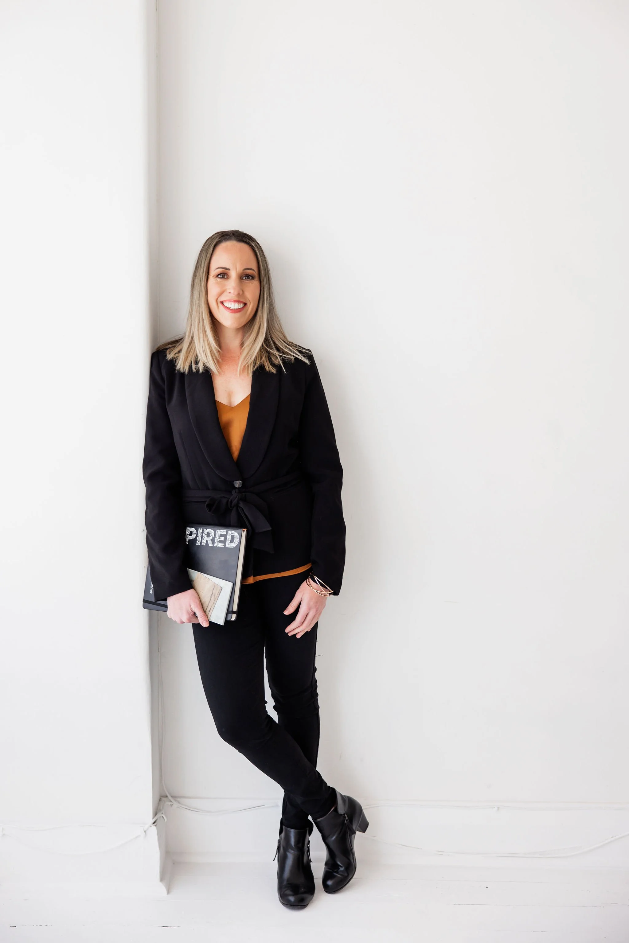 A woman standing against a white wall, holding a notebook titled "PIRED", wearing a black blazer, black pants, and black boots, smiling at the camera.