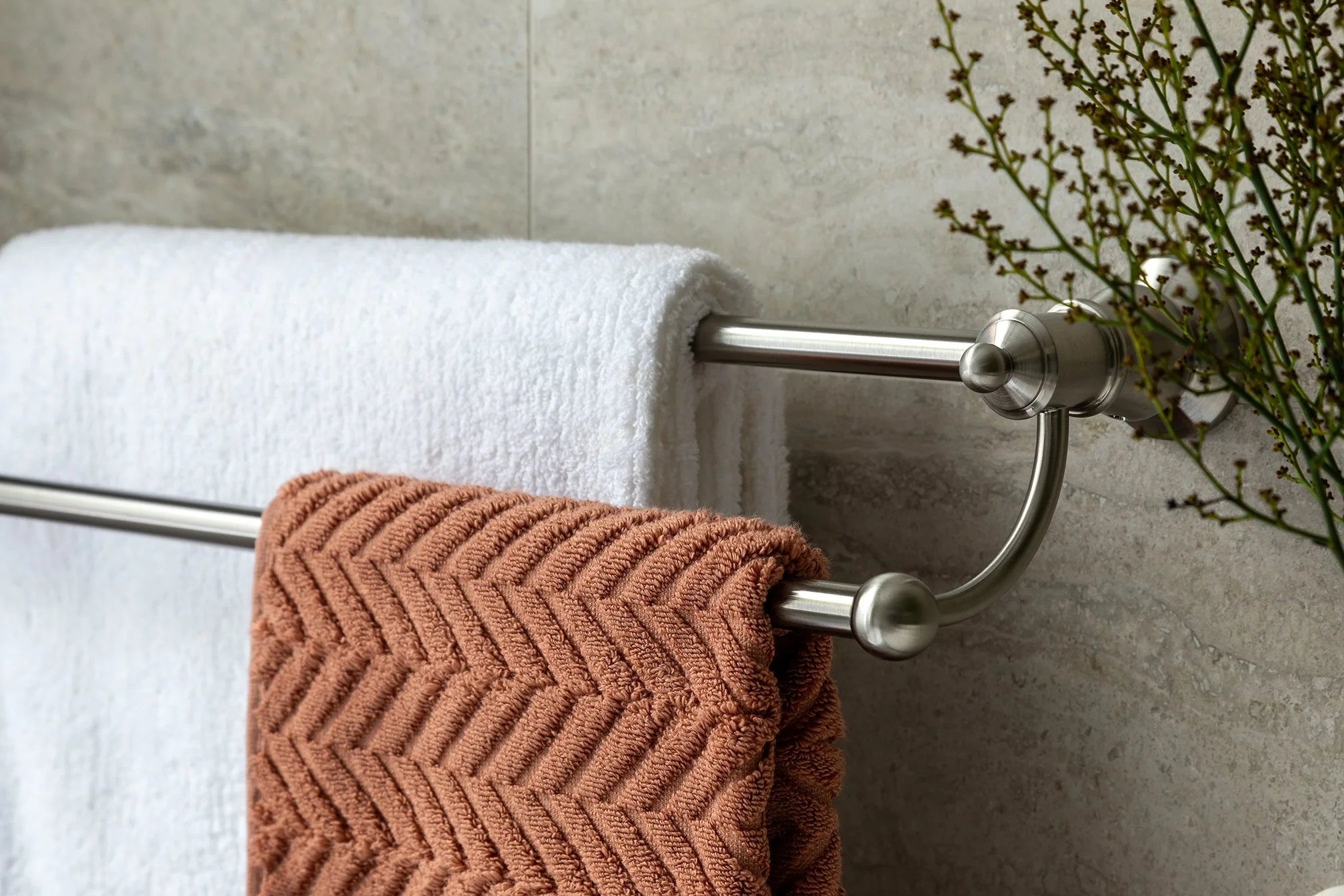 A metal towel rack mounted on a beige tiled wall holding a white towel and a rust-colored textured towel. Green plant branches are visible on the right side.