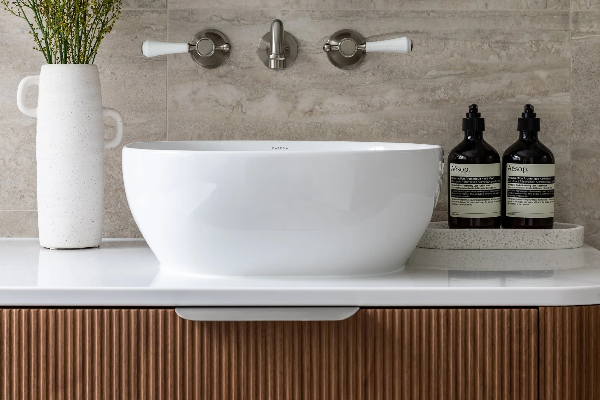Bathroom sink with two soap dispensers on a tray, a tall white vase with greenery, and a modern wall-mounted faucet against beige tiled wall.