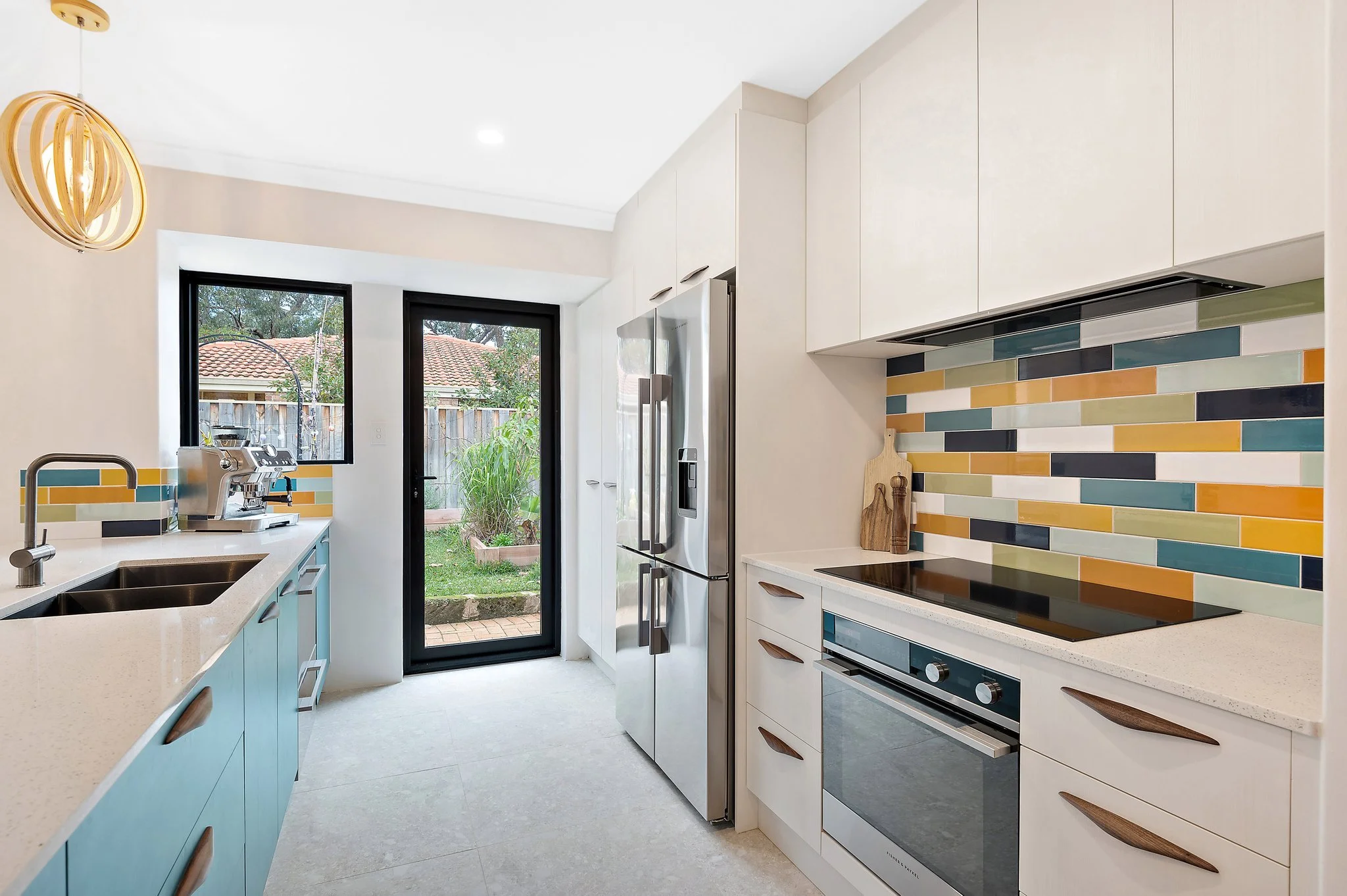 Modern kitchen with multicolored backsplash, stainless steel refrigerator, electric stove, and light-colored cabinetry. There is a door leading outside to a grassy yard.