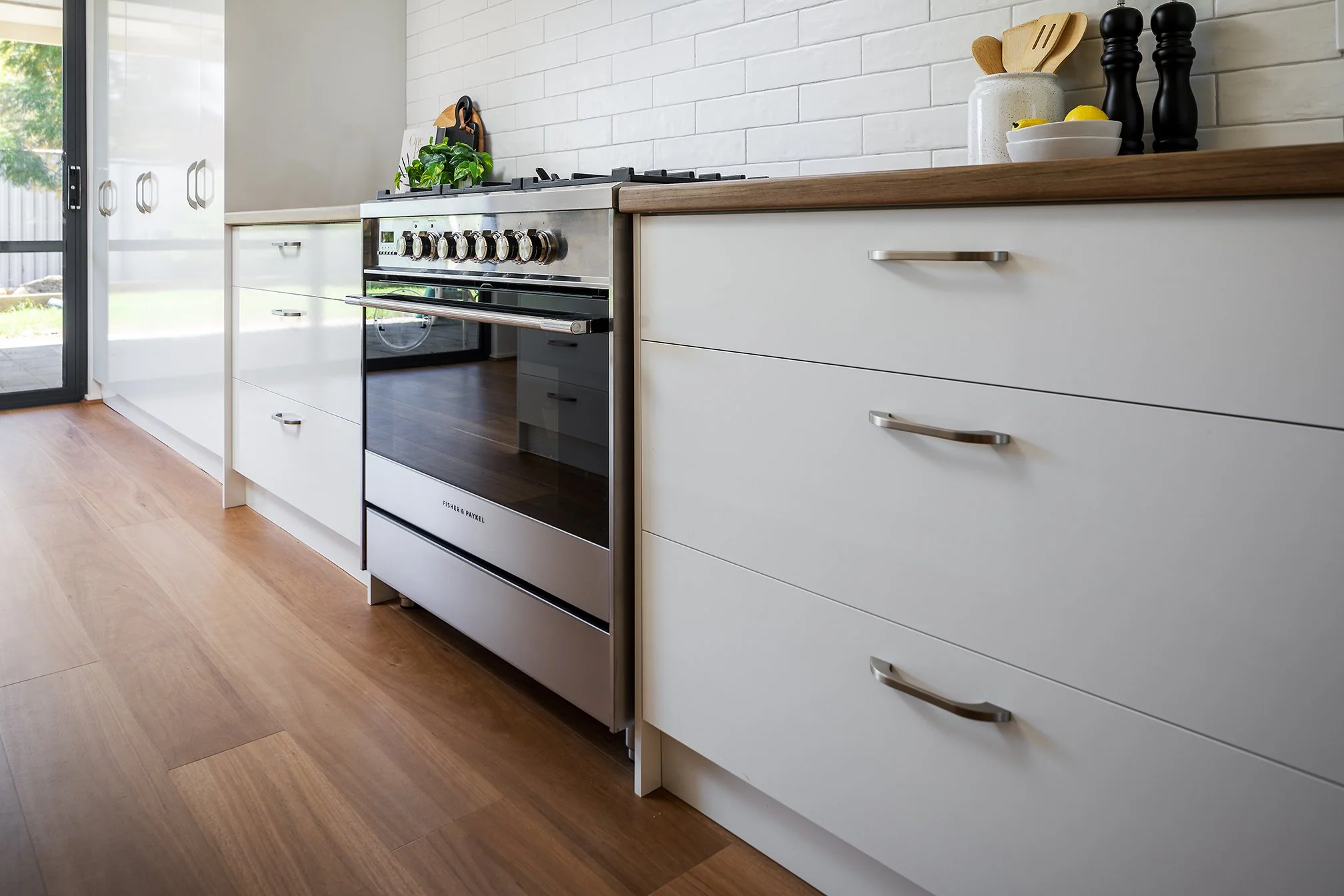 Modern kitchen with white cabinetry, a stainless steel oven, and a wooden countertop. There are potted plants, jars, and utensils on the counter, and a sliding glass door leading outside.
