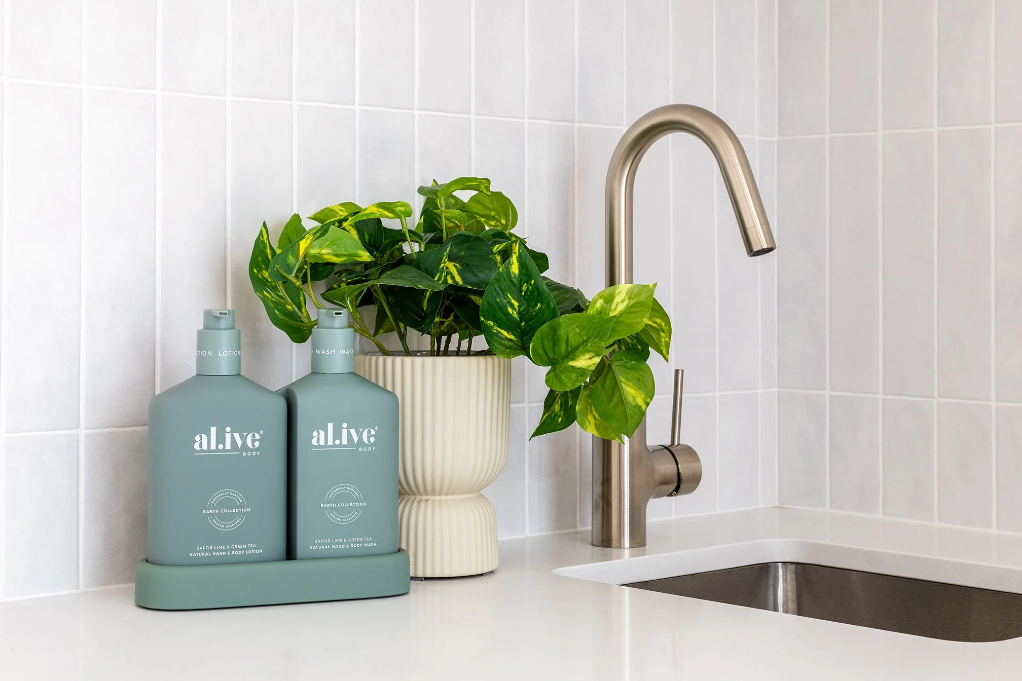 Kitchen sink area with two bottles of a.livé body lotion and wash, a potted green plant, stainless steel faucet, white tiled backsplash, and a white countertop.