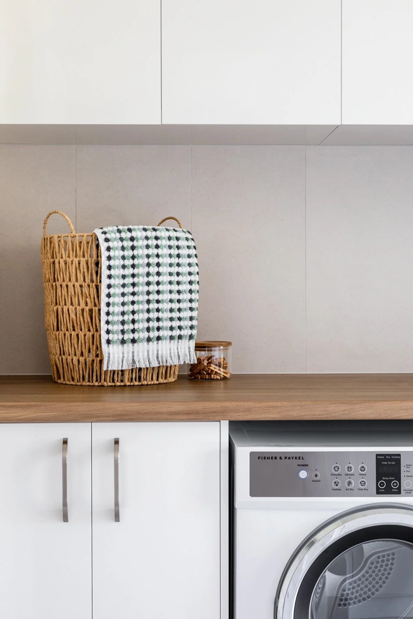A laundry room with white cabinets, a wooden countertop, and a washing machine. There is a wicker laundry basket with a patterned towel hanging over it, and a small glass jar with clothespins on top.
