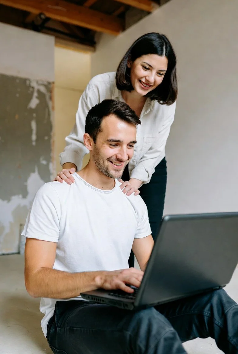 A woman and a man smiling and looking at a laptop, sitting on the floor in a room with unfinished walls and exposed wooden ceiling beams.