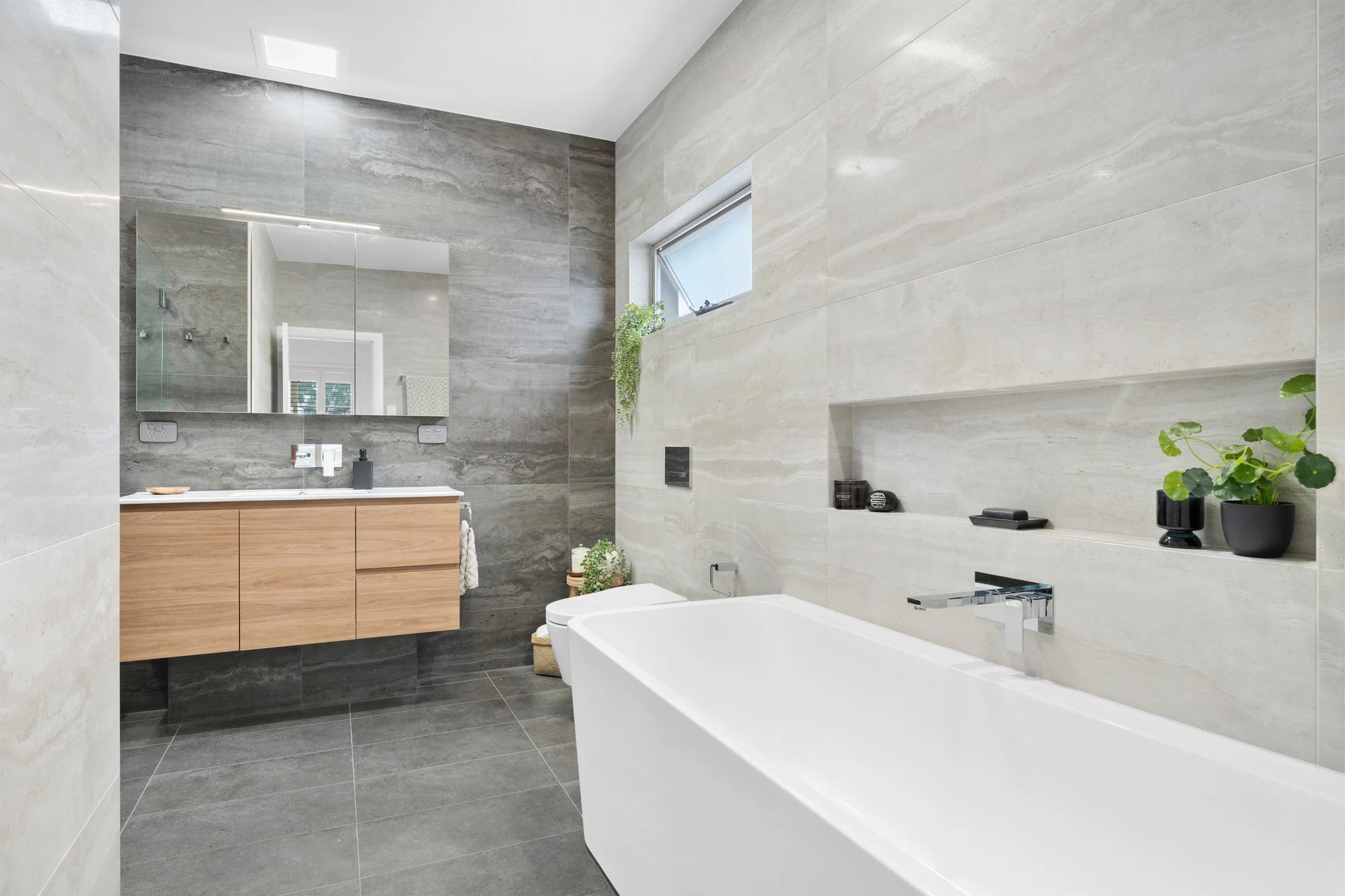 Modern bathroom with a freestanding bathtub, a large mirror, a wooden vanity, a window with plants, and neutral-colored tiles.