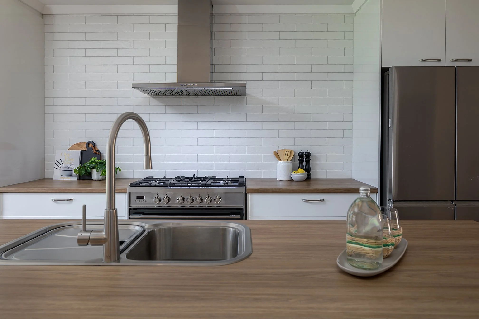 Modern kitchen with a countertop, sink, and faucet in the foreground, a stove with an exhaust fan, and a large refrigerator in the background. Decor includes a small plant, a cookbook, a jar with utensils, and a tray with water bottles and glasses.