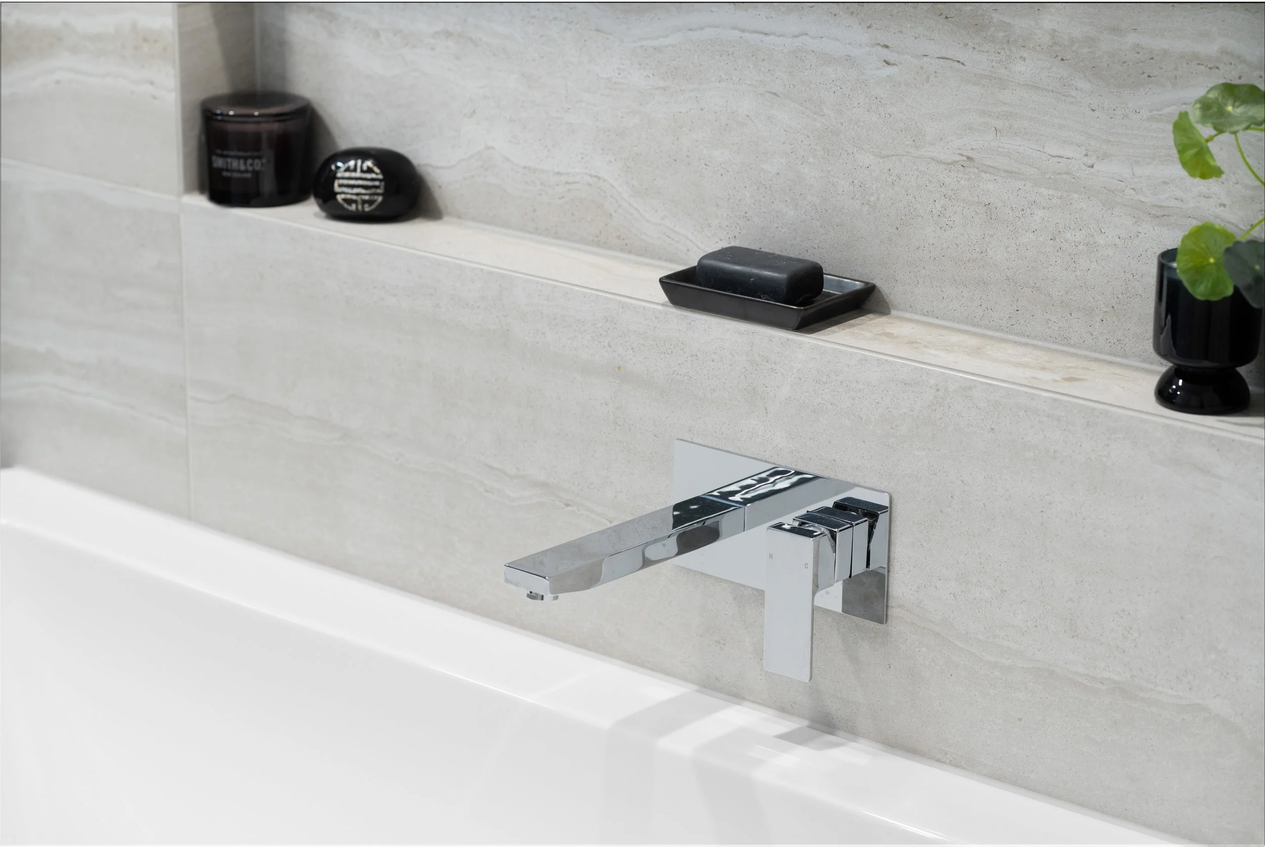 Close-up of a modern bathroom faucet mounted on a light beige tiled wall, with a shelf above holding black containers and a soap dish with a black soap bar, and part of a white bathtub.