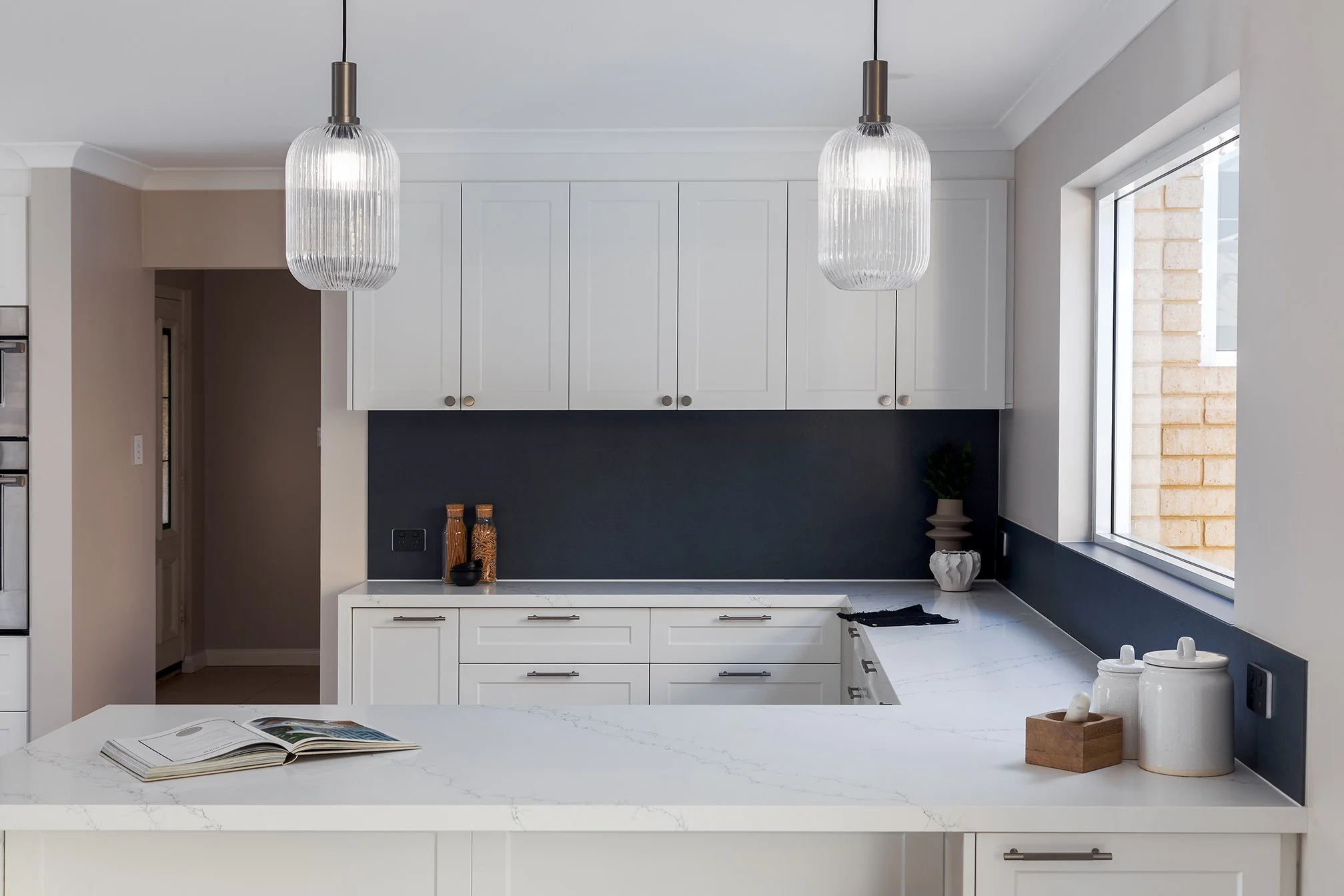 Modern kitchen with white cabinets, dark backsplash, marble countertop, large window, and two hanging pendant lights.