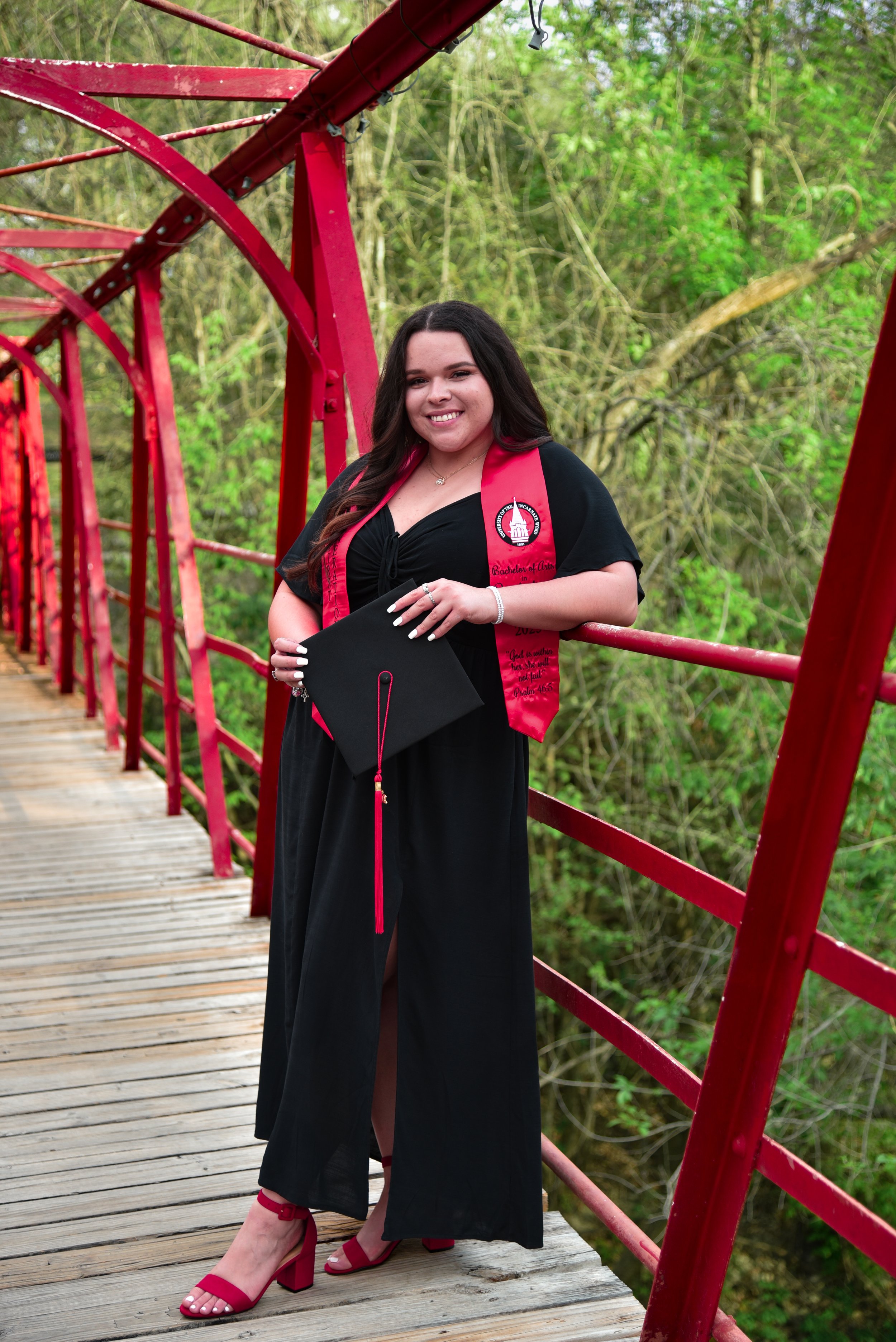 A young woman in a black graduation gown and red sash standing on a wooden bridge with red railing, holding a graduation cap, smiling, with green trees in the background.