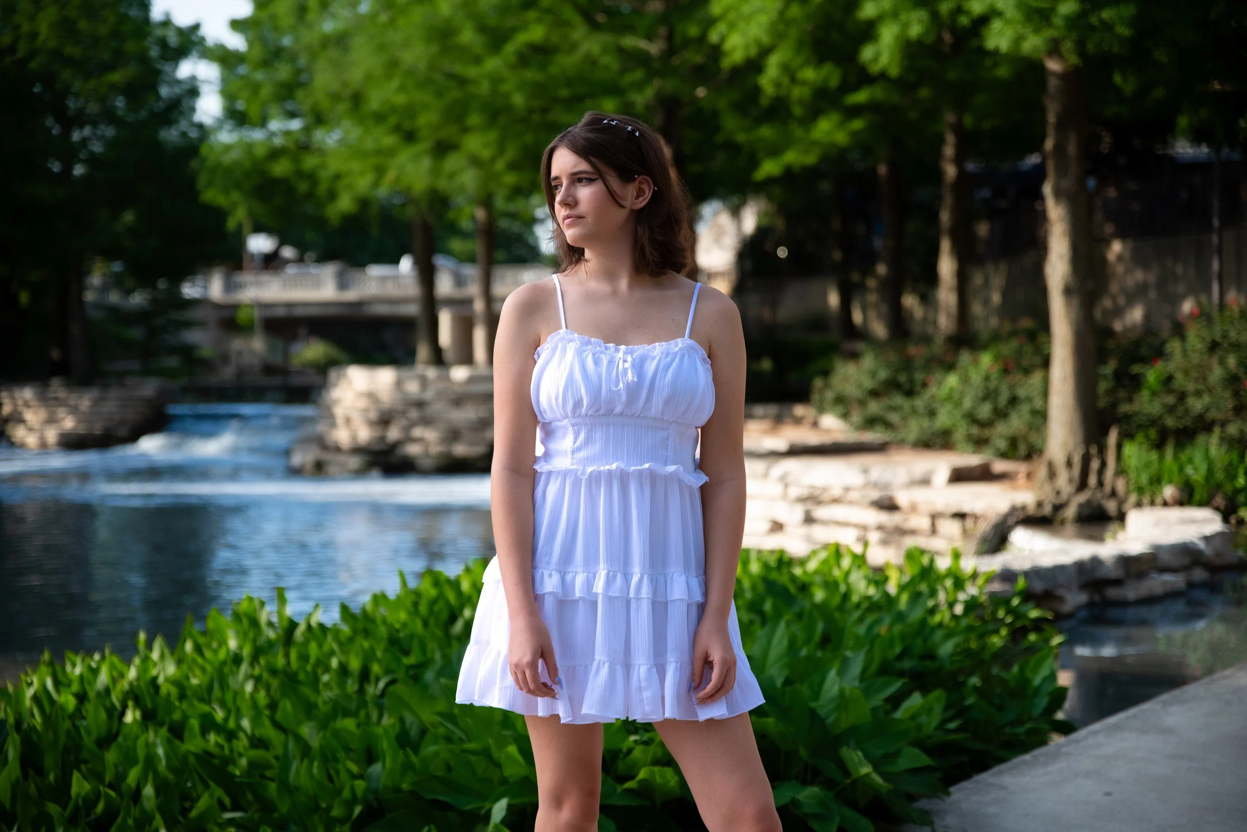 Young woman in white dress standing outdoors near water with trees and greenery in background.