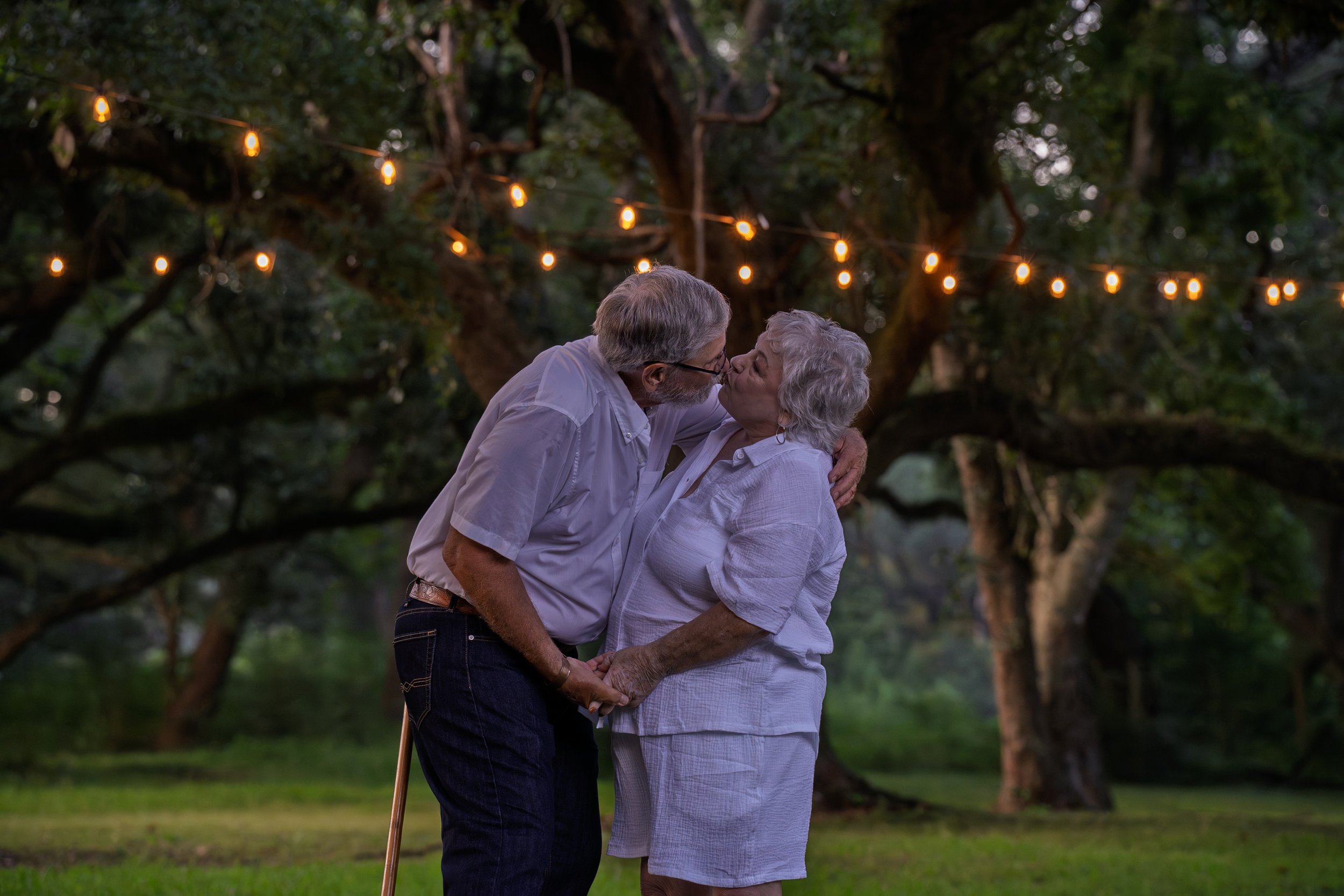 An elderly couple kissing outdoors under string lights, with large trees in the background during evening.