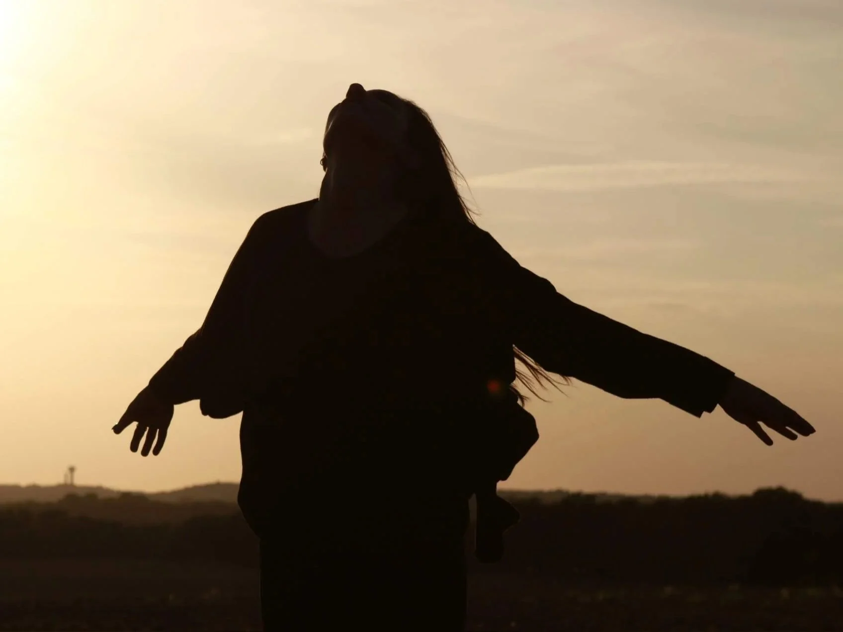 Silhouette of a woman with outstretched arms standing outdoors during sunset or sunrise.
