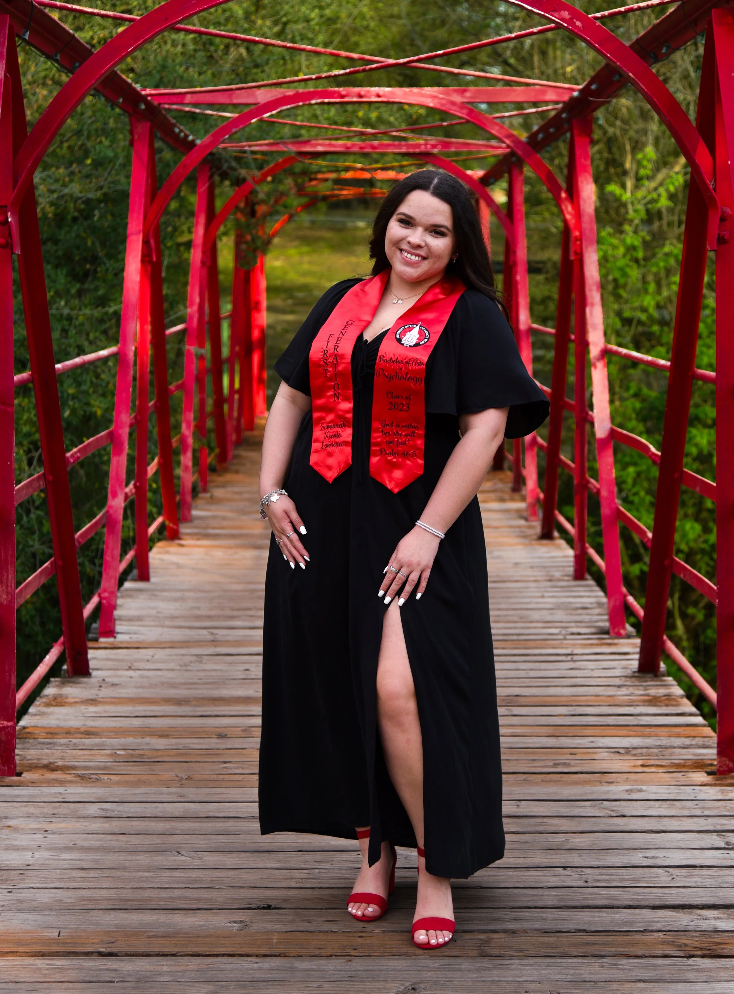 A young woman in a graduation gown on a wooden bridge, wearing a red stole with academic and scriptural text, smiling at the camera.