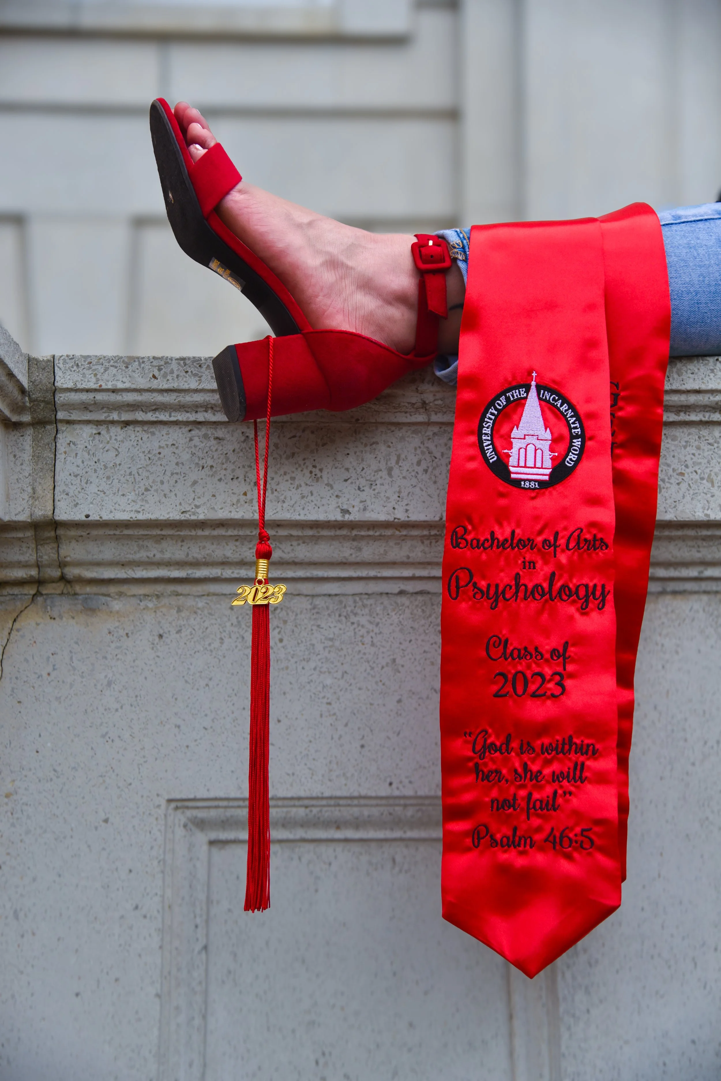 Graduation cap and stole draped over a stone ledge, with a shoe resting on top. The stole reads 'Bachelor of Arts in Psychology Class of 2023' and features a quote from Psalm 46:5.