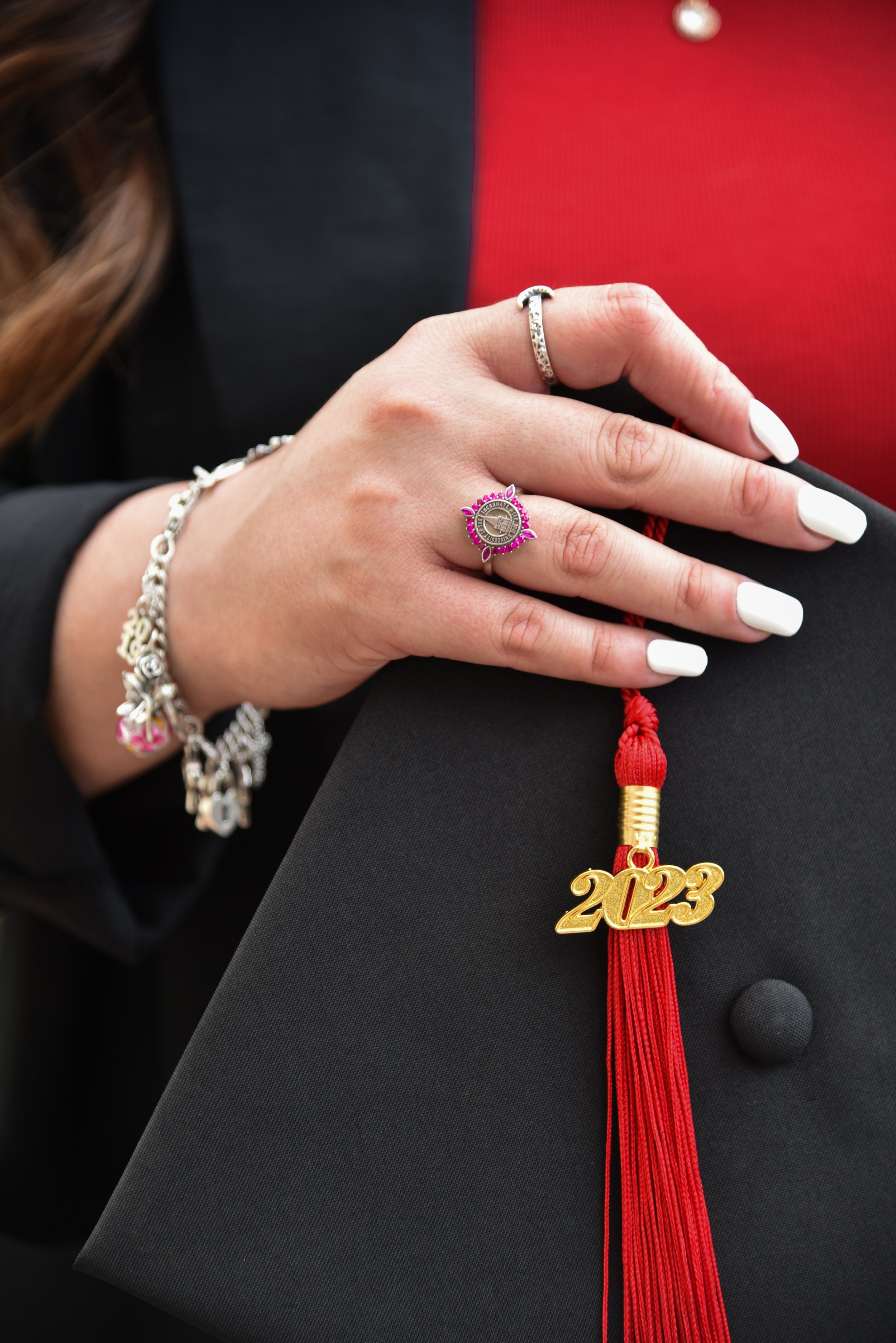 Close-up of a woman's hand resting on a graduation cap with a red tassel that has a "2023" charm. She is wearing multiple rings, including a silver ring with black stones, a pink-bordered round charm with a silver design, and a silver bracelet with pink accents. Her nails are painted white.