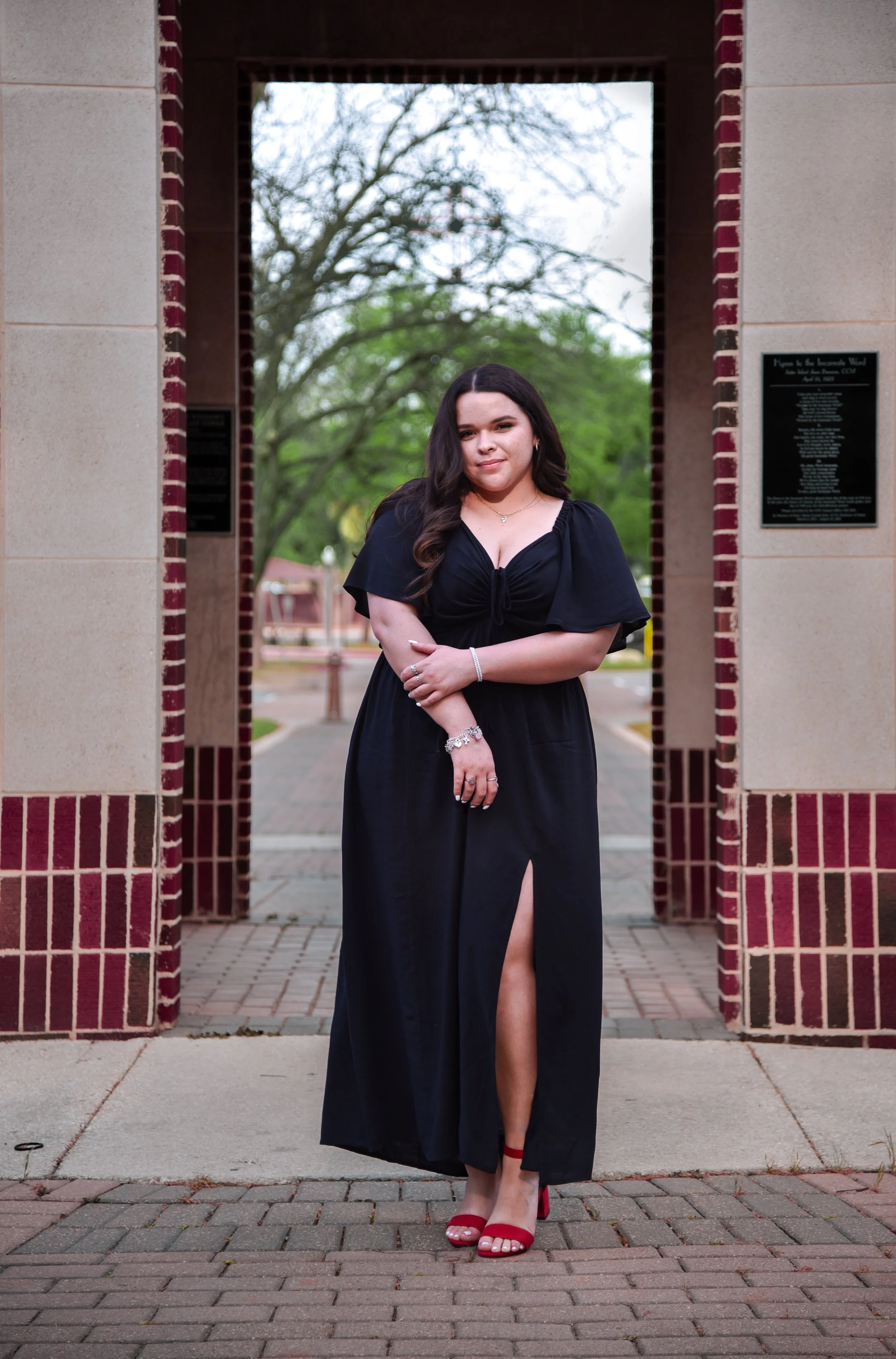 Young woman in a black dress standing in an archway on a sidewalk, with trees and a park in the background.