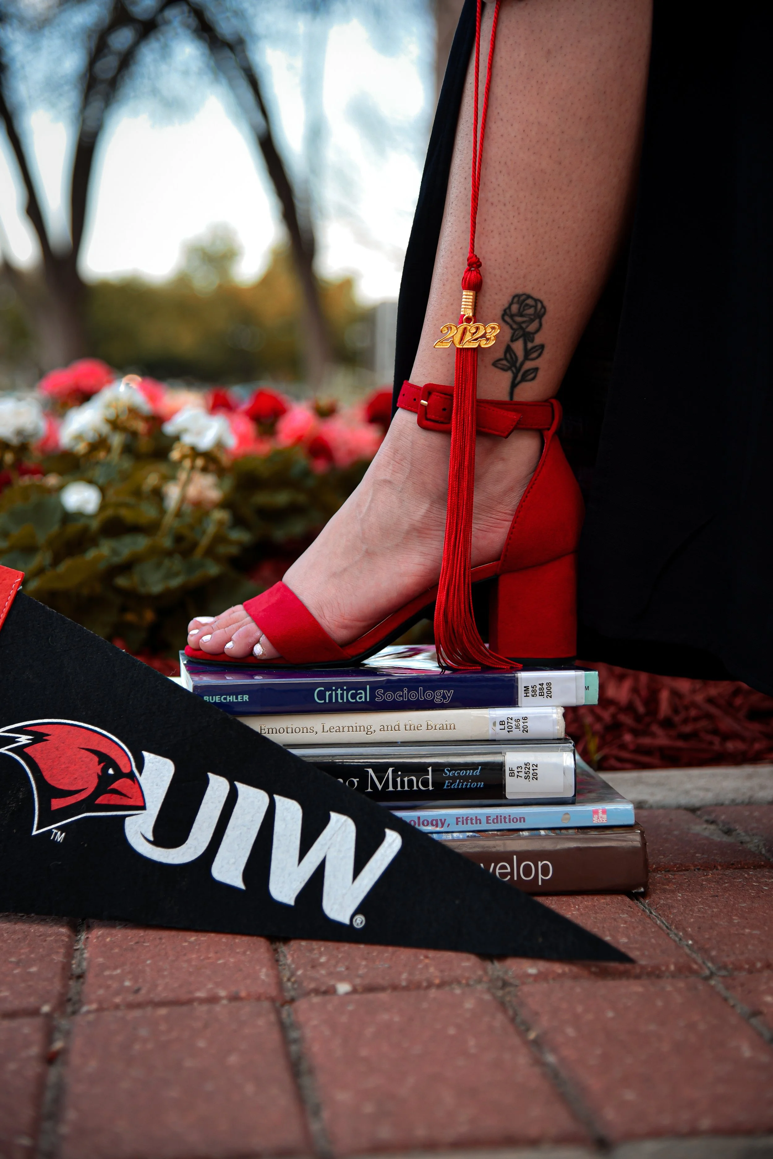 Close-up of a person's foot on a stack of textbooks on a brick sidewalk. The person is wearing a red high-heeled sandal, a red ankle strap, and a red tassel with a '2023' charm. The person has a tattoo of a rose on their ankle. The textbooks include titles such as 'Critical Sociology,' 'Emotions, Learning, and the Brain,' and 'Psychology, Fifth Edition.' Part of a black and orange University of Wisconsin-Milwaukee pennant is visible in the foreground, and background includes trees and blurred foliage.