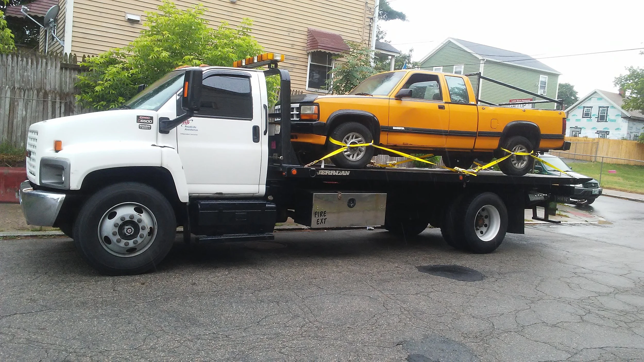 A white tow truck carrying a yellow pickup truck secured with yellow straps on its flatbed, parked on a street in front of residential houses.