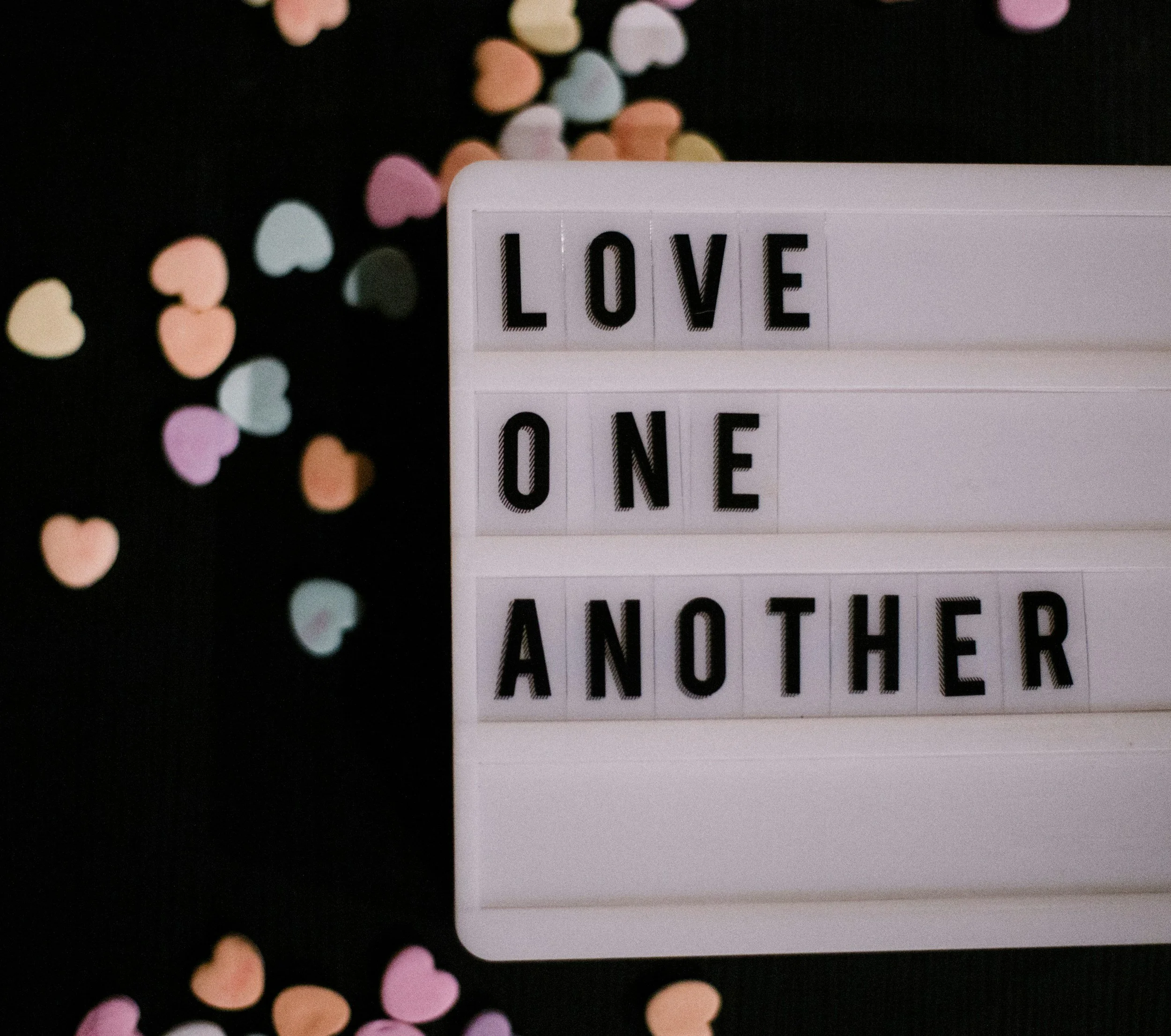 A lightbox with black letters on a white background spelling out 'LOVE ONE ANOTHER' surrounded by heart-shaped confetti in pastel colors on a black surface.