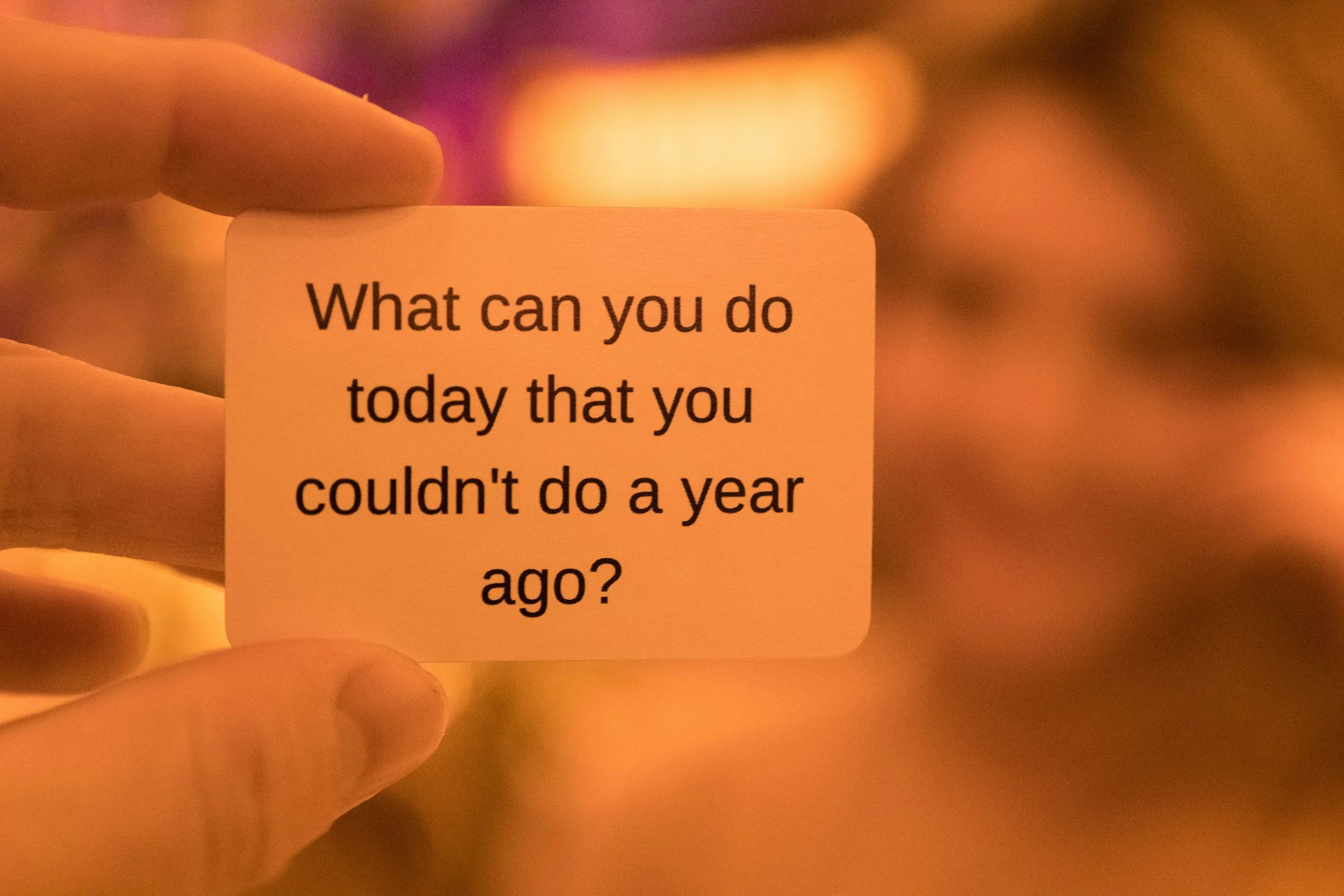 A hand holding a small card with the text 'What can you do today that you couldn't do a year ago?' written on it, with colorful, blurred lights in the background.