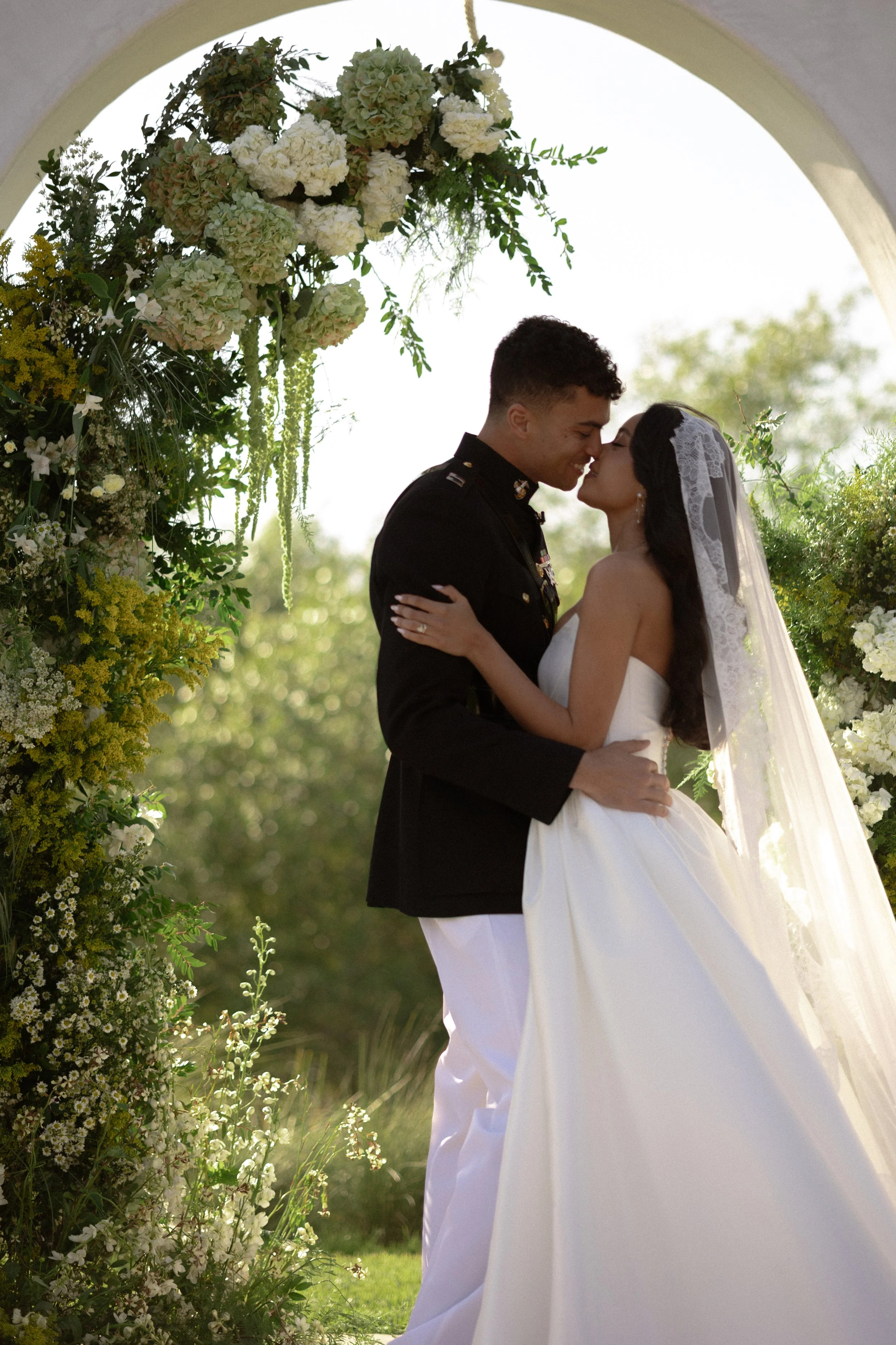 A newlywed couple sharing a kiss under a floral wedding arch outdoors in bright daylight.