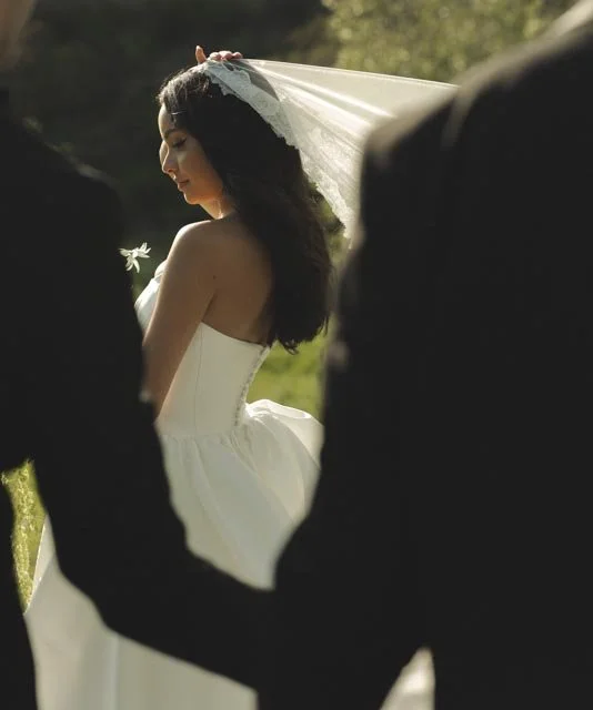 A bride with long dark hair, wearing a strapless white wedding dress and veil, holds a flower while standing outdoors during the daytime.