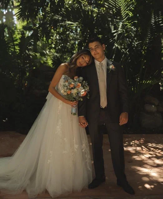 Bride and groom holding hands outdoors with green foliage background, bride in white wedding gown holding a bouquet, groom in black suit and tie.