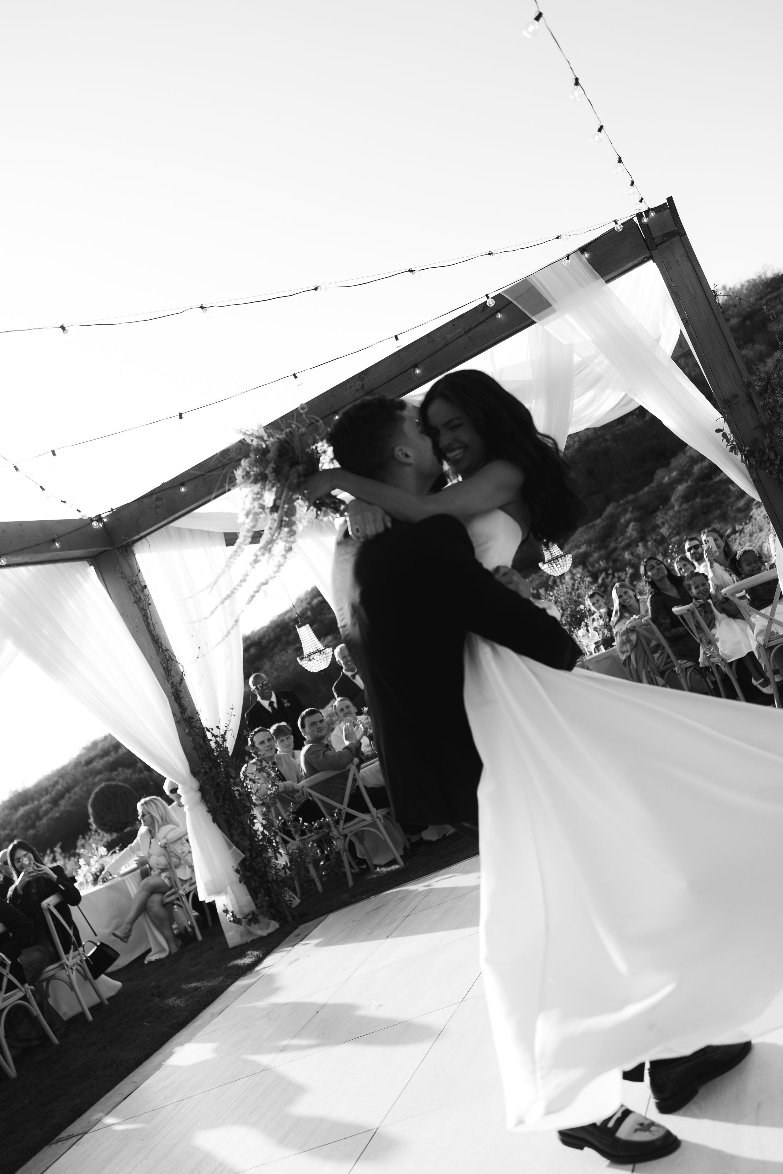 A black and white photo of a newlywed couple dancing at their outdoor wedding reception, with guests seated at tables in the background, under a decorated wooden arch with curtains and string lights.