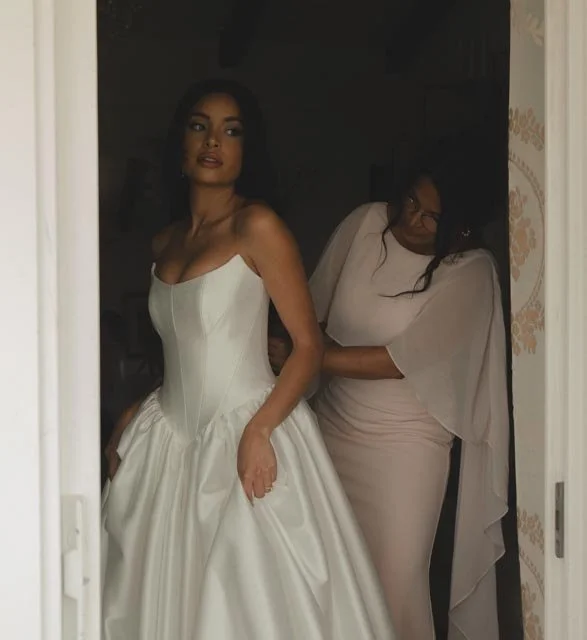 A woman in a white wedding gown standing in a doorway, with another woman in a light pink dress adjusting her dress.