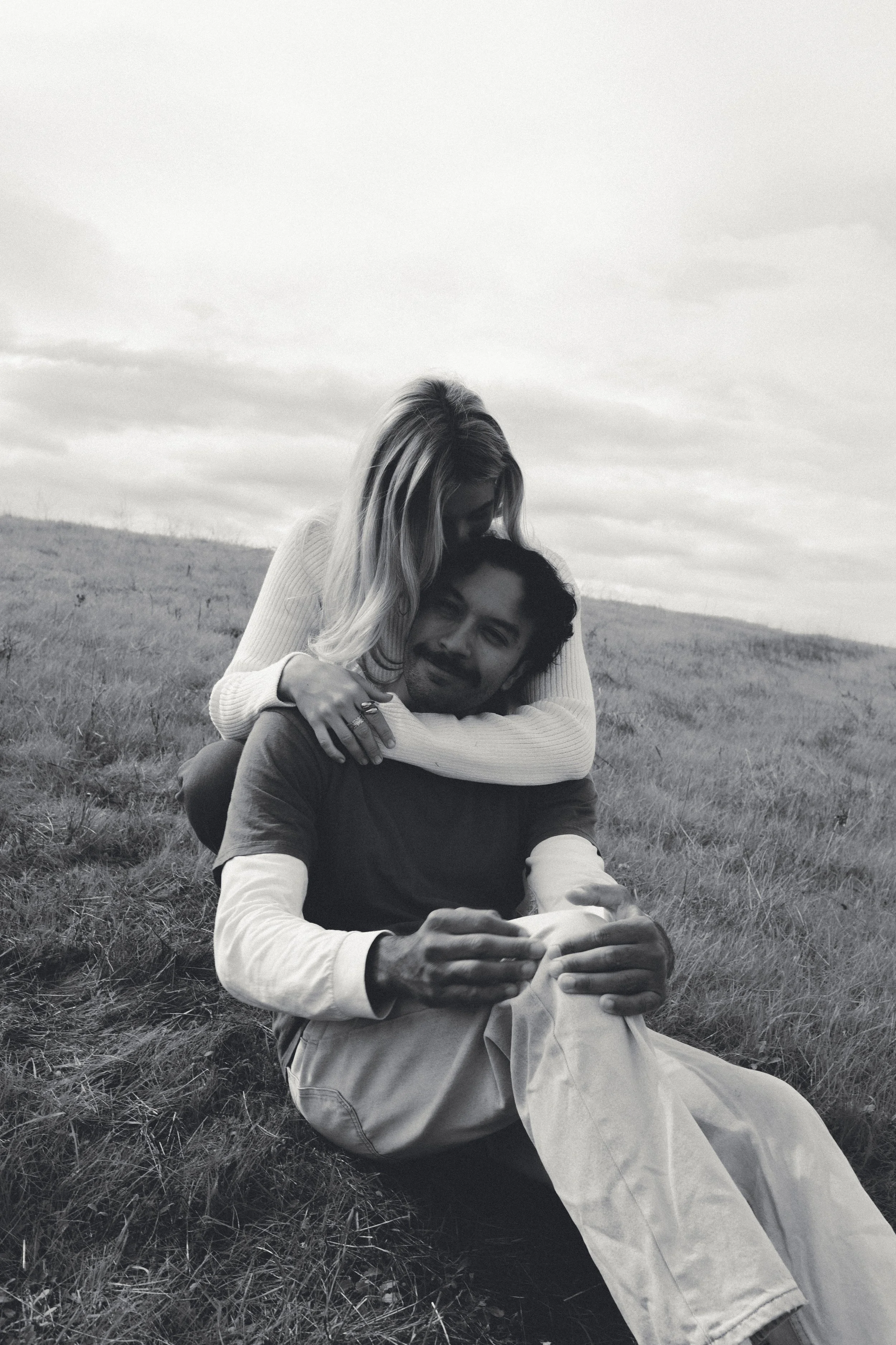 A black-and-white photo of a woman hugging a man while sitting on grass in an open field under a cloudy sky.