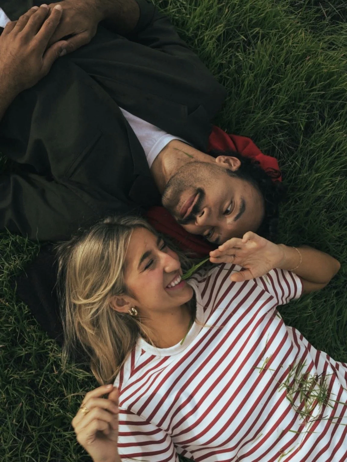 A man and woman lying on grass, smiling and playing with a blade of grass.