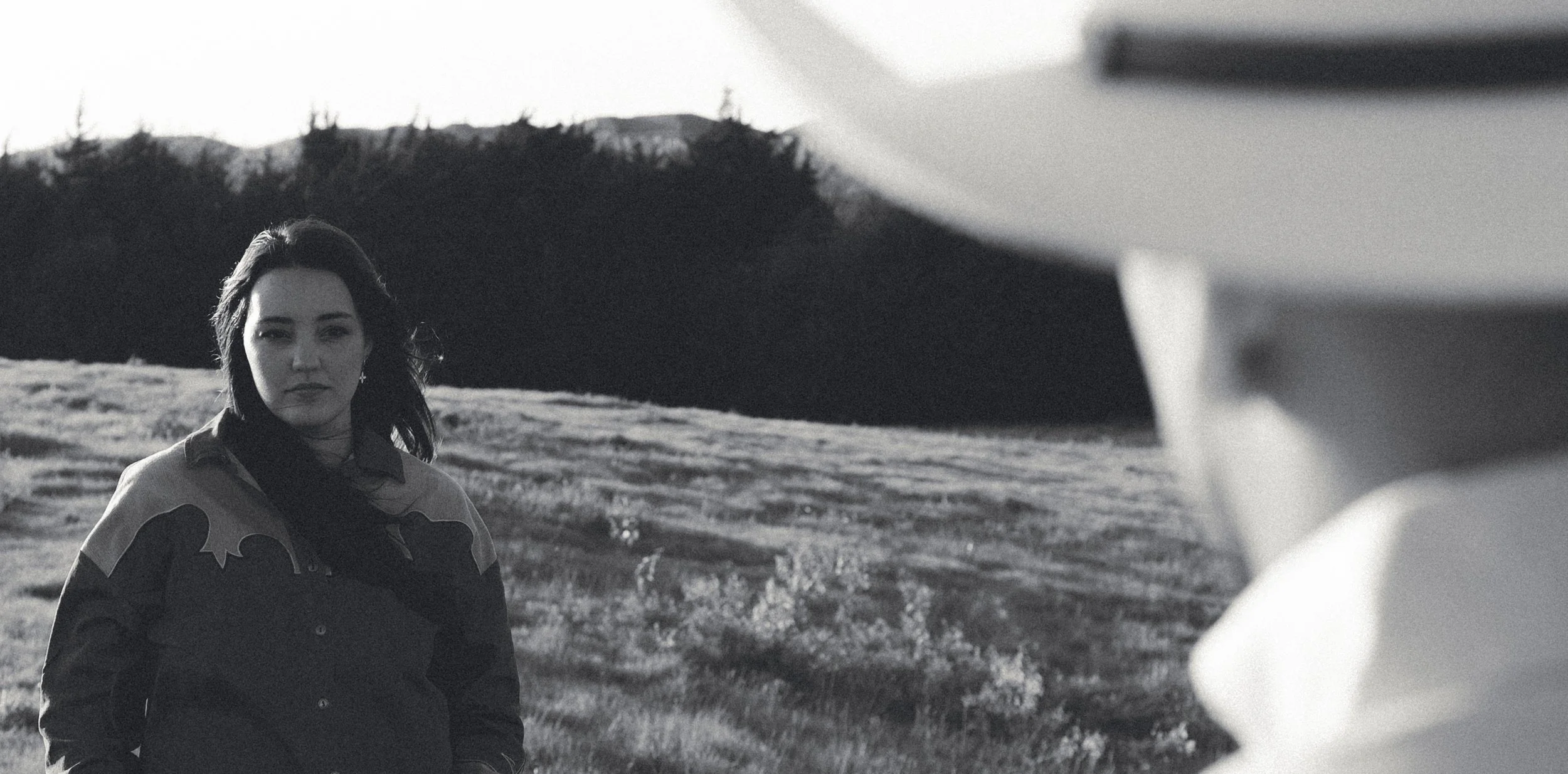 A woman with dark hair wearing a jacket stands outdoors in a grassy field with hills and trees in the background. The image is in black and white.