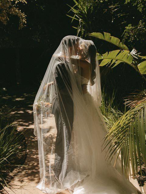 Bride in a wedding dress stands outdoors with a veil covering her face, surrounded by lush green tropical plants.